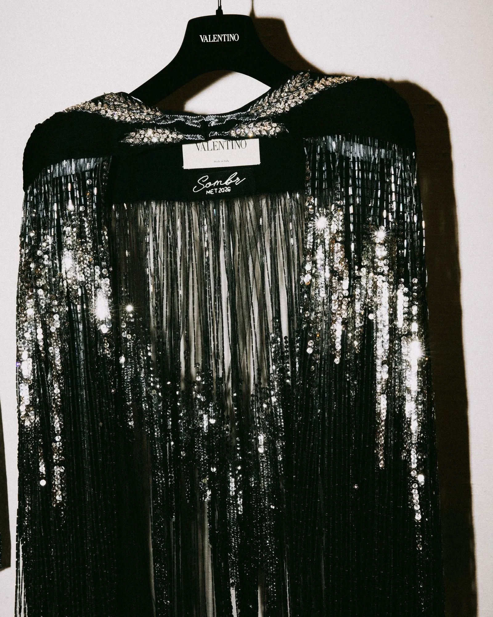 Black sequined dress hanging on a Valentino branded hanger, with metallic fringe details visible on the bodice and skirt edges.