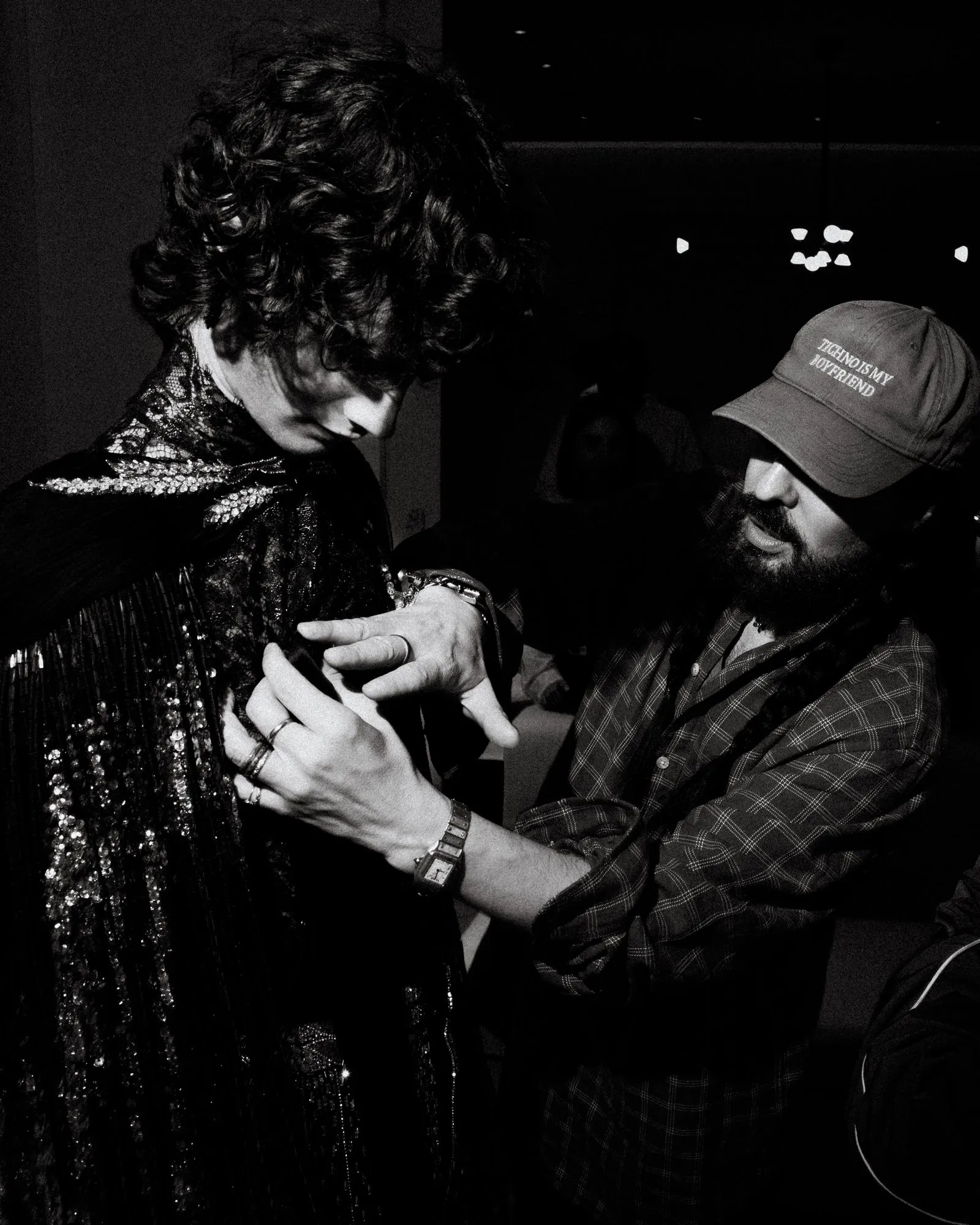 Close-up of two people shaking hands; left person in a shiny sequined dress, right person in a plaid shirt and cap.