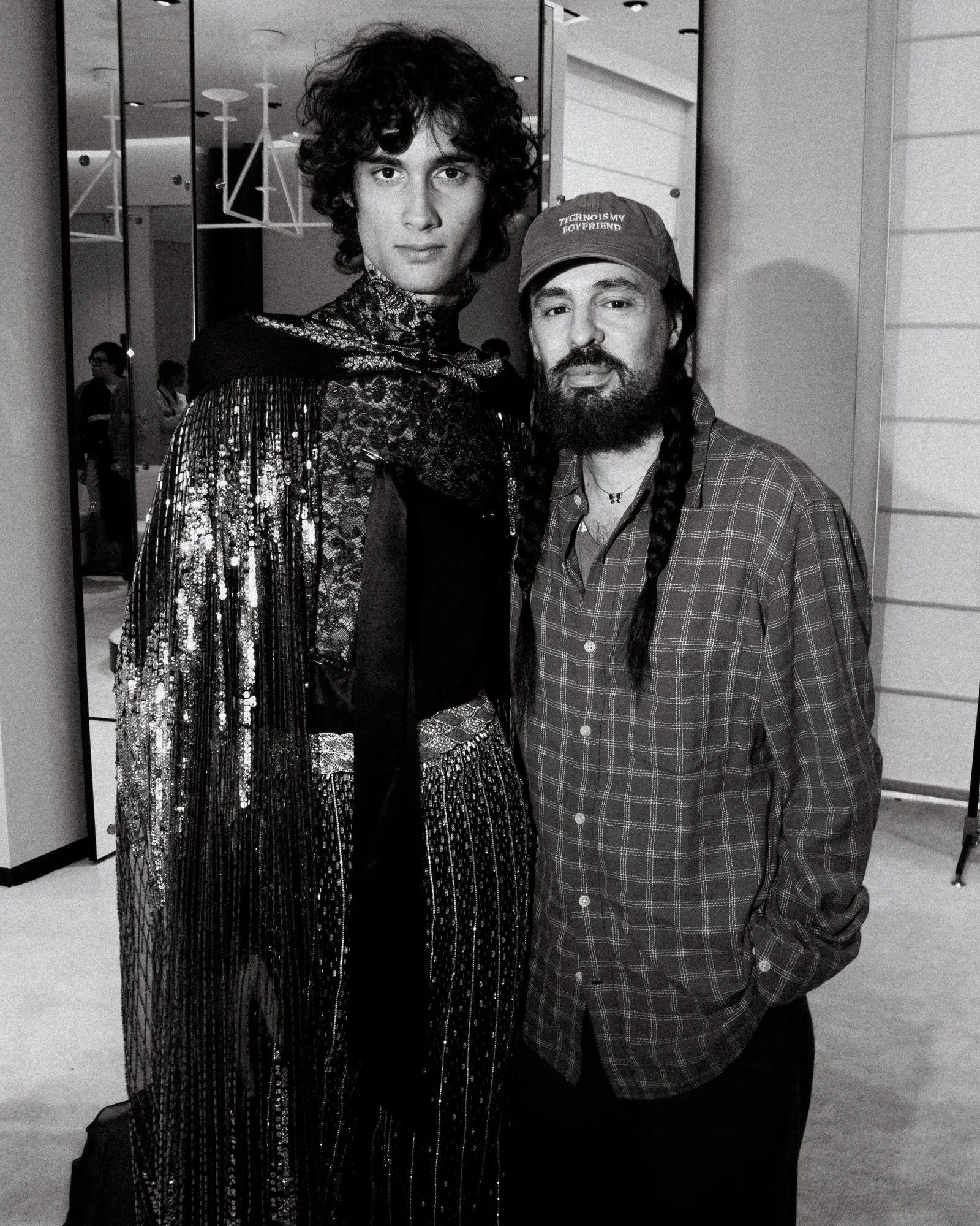 Two people stand close together indoors; one wears a shimmering lace dress with sequins, the other wears a plaid shirt and a cap, posing for the photo.