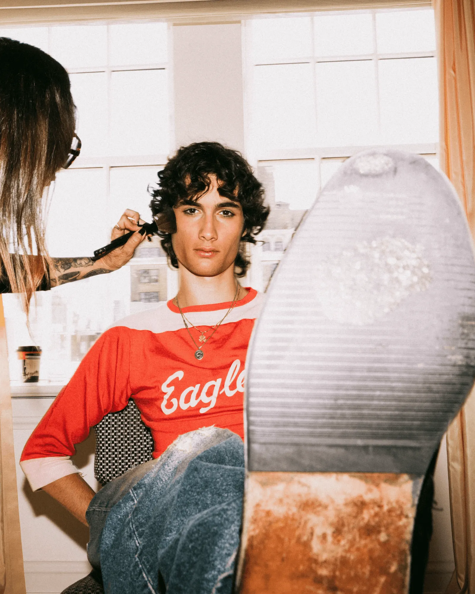 Young man with curly dark hair is getting makeup applied to his face by a makeup artist with a tattooed forearm, wearing a red shirt and jeans in a bright room.