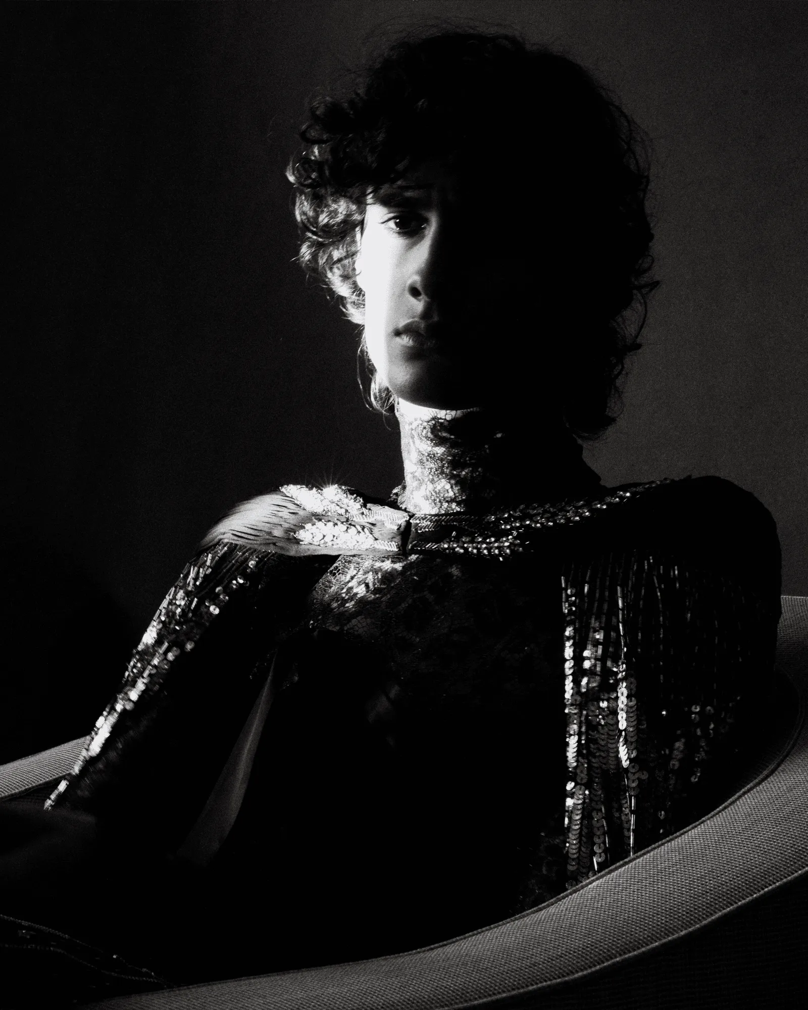 High-contrast black-and-white portrait of a person with half the face in shadow, wearing a sequined neckline and ornate collar.