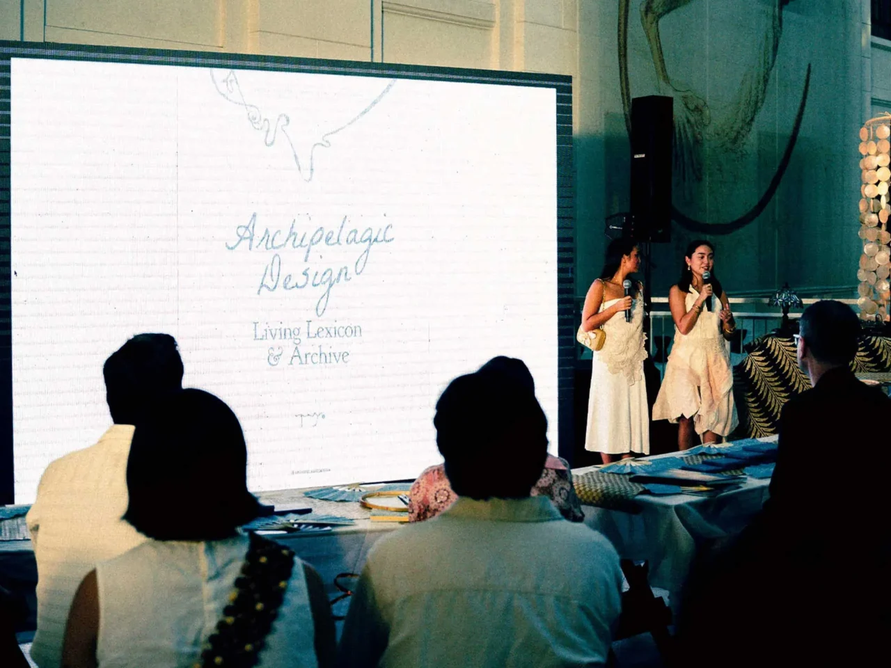 Two women in white dresses speak into microphones on a stage beside a large screen that reads 'Archipelago Design' and 'Living Lexicon & Archive'.