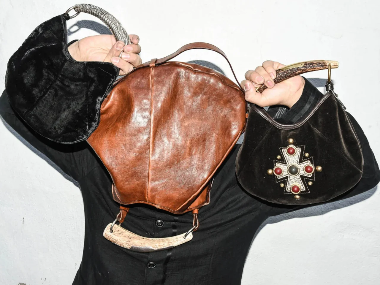Person in a black robe grips a brown leather backpack with a curved wooden handle against a light background, palms visible near the bag’s strap.