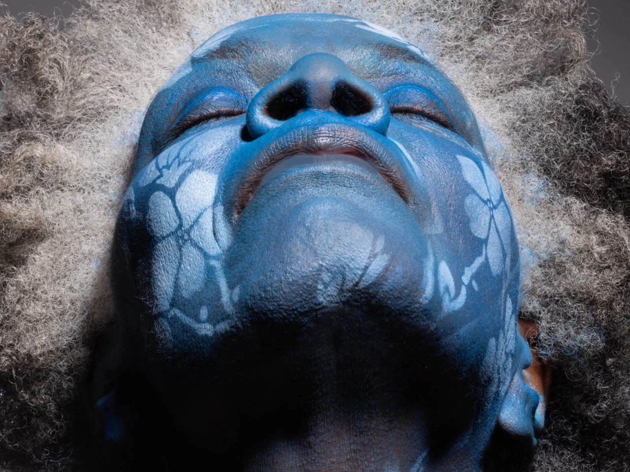 Close-up of a person with blue face paint and curly gray hair, eyes closed, lying on their back.