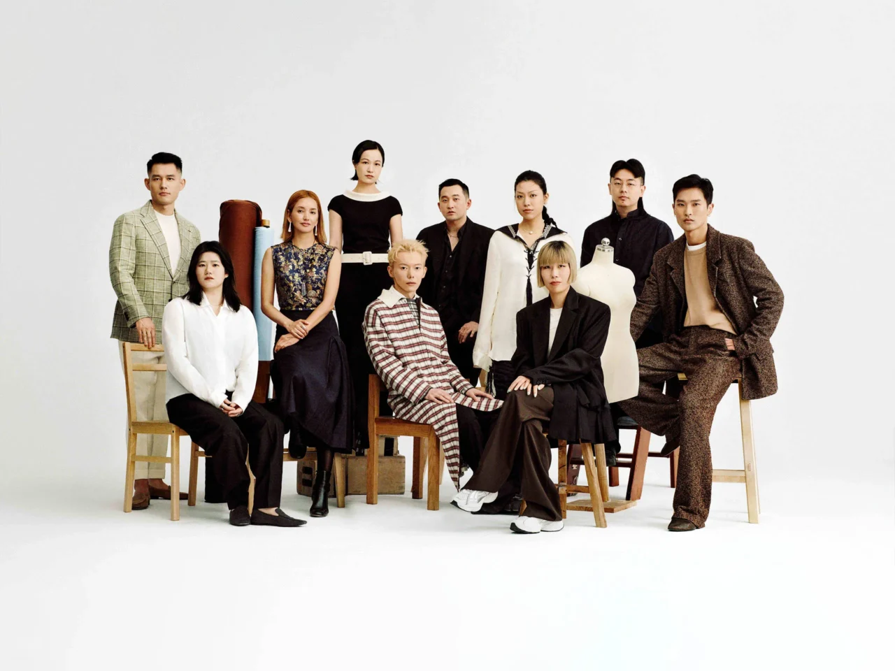 Group of ten fashion models posing in a studio against a white backdrop, wearing diverse outfits and sitting or standing with stools and mannequins nearby.