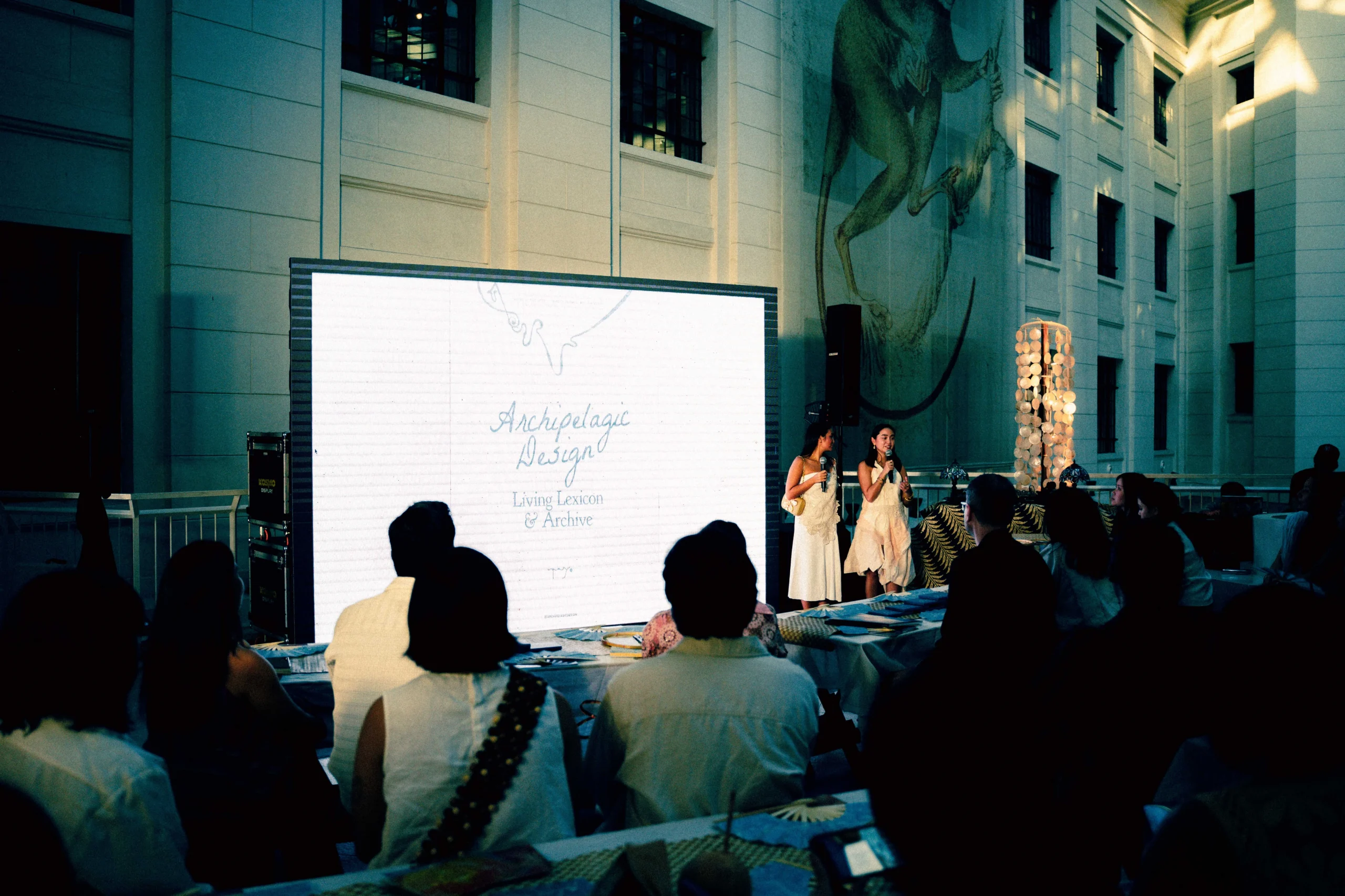 Two women in white dresses speak into microphones on a stage with a large illuminated screen behind them showing Archipelago Design text and a logo.