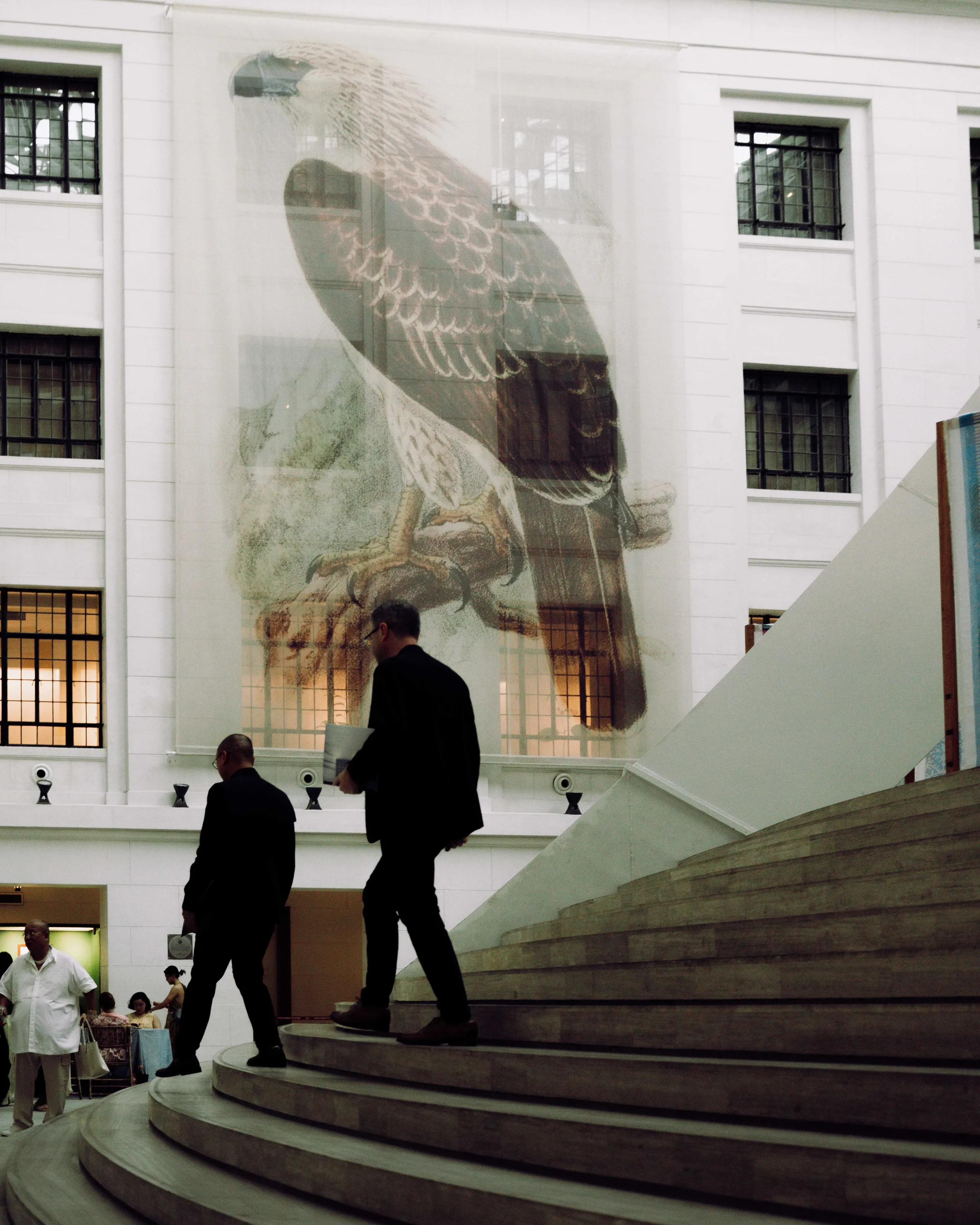 Large glass wall mural of a bird (eagle) over a white modern atrium with curved stone steps and people walking byshi