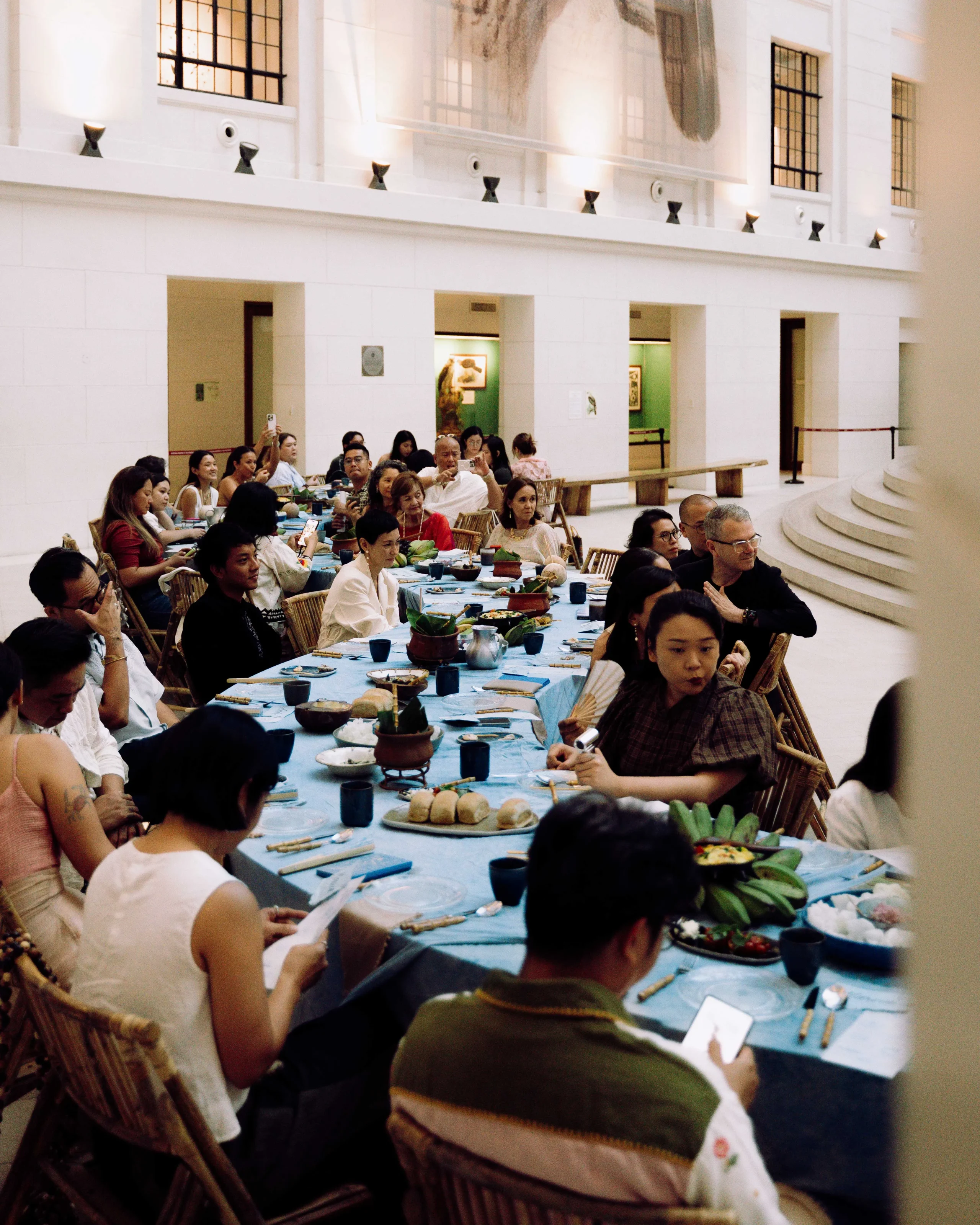 Large group of people seated along a long banquet table in a bright, white-walled hall, eating and socializing.
