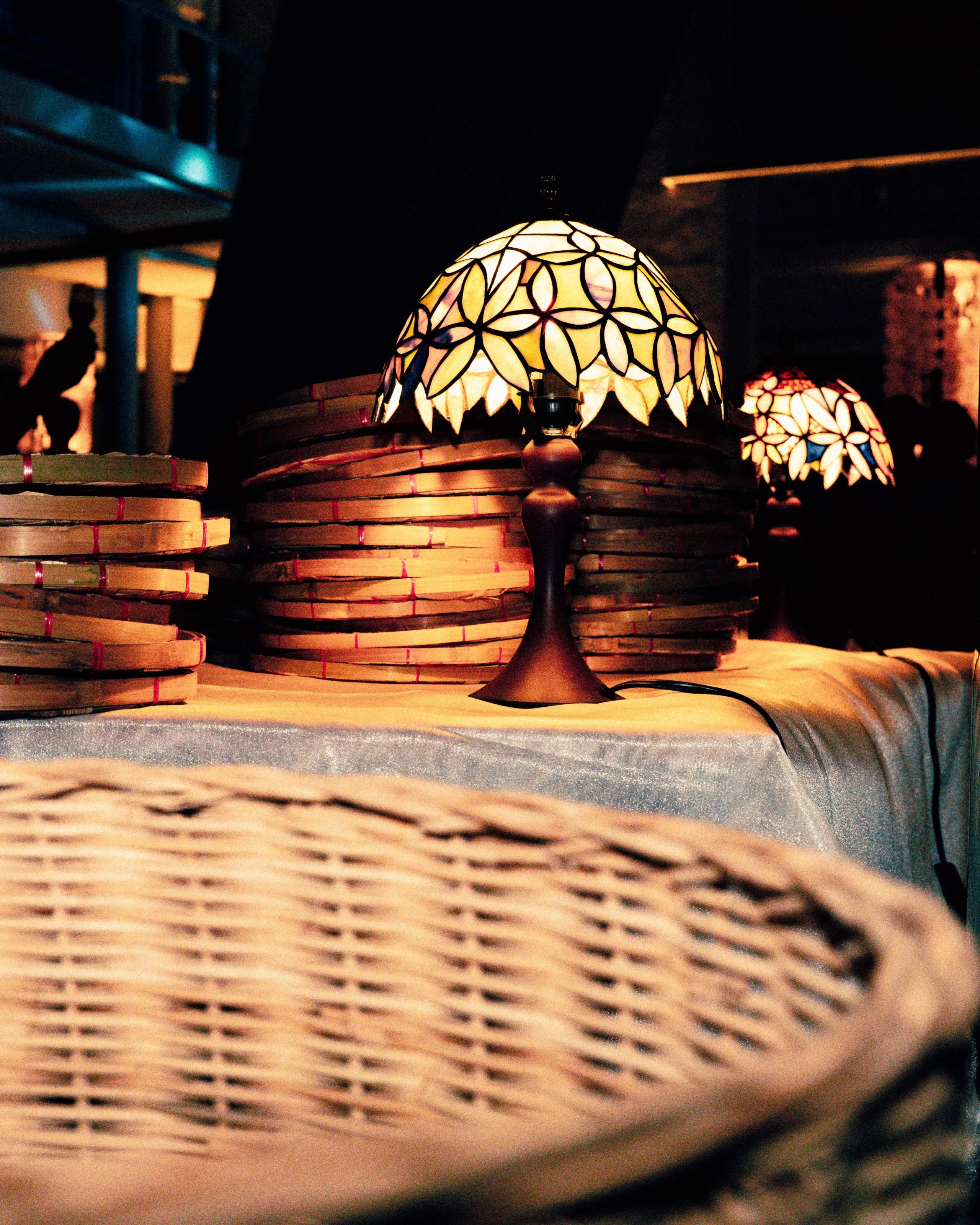 Tiffany-style lamp with a colorful stained-glass shade lit, placed among stacked wicker baskets in a dim setting.