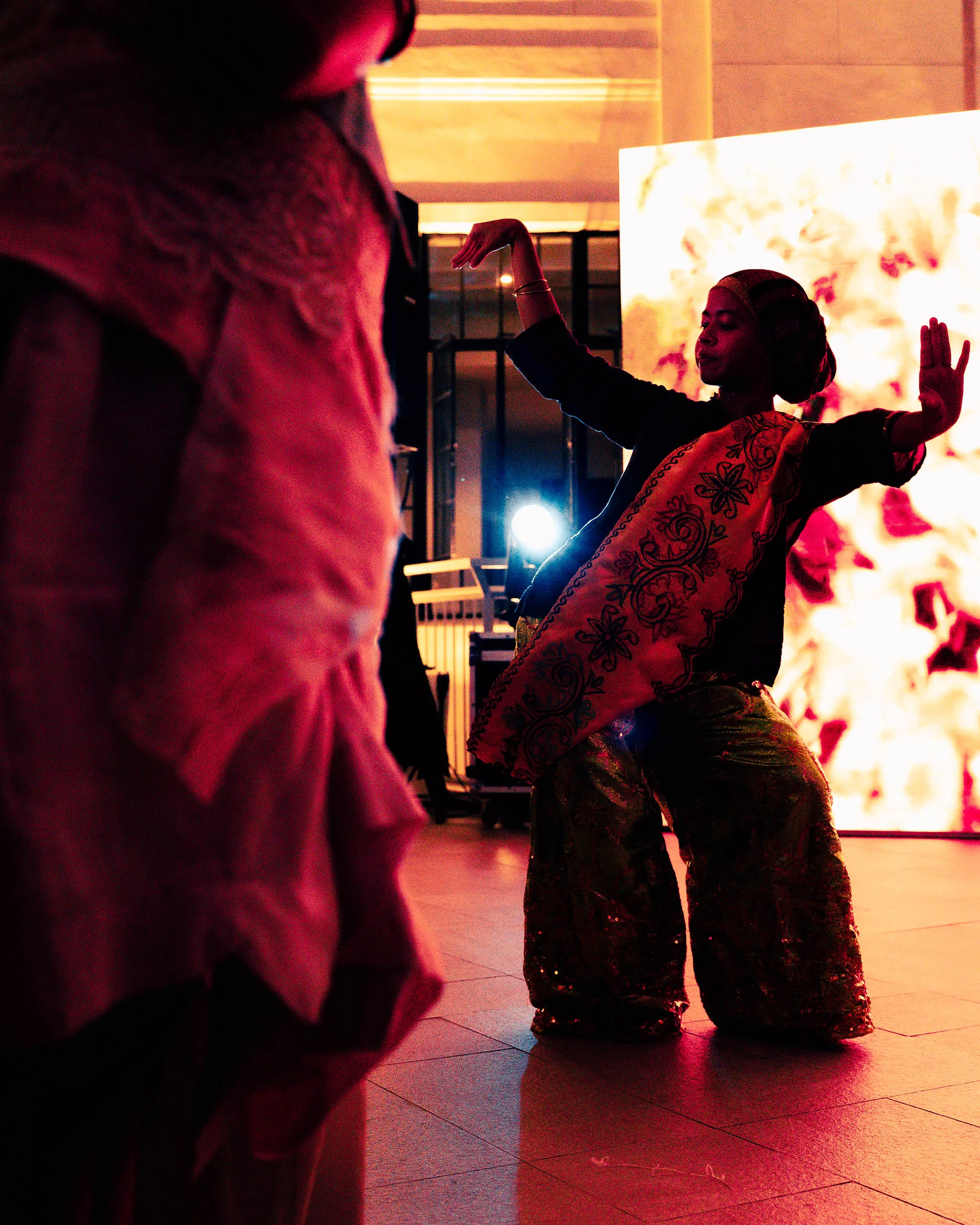 Dancer in a traditional patterned dress with arms raised on a warmly lit stage, foreground silhouette of another performer in the left foreground.