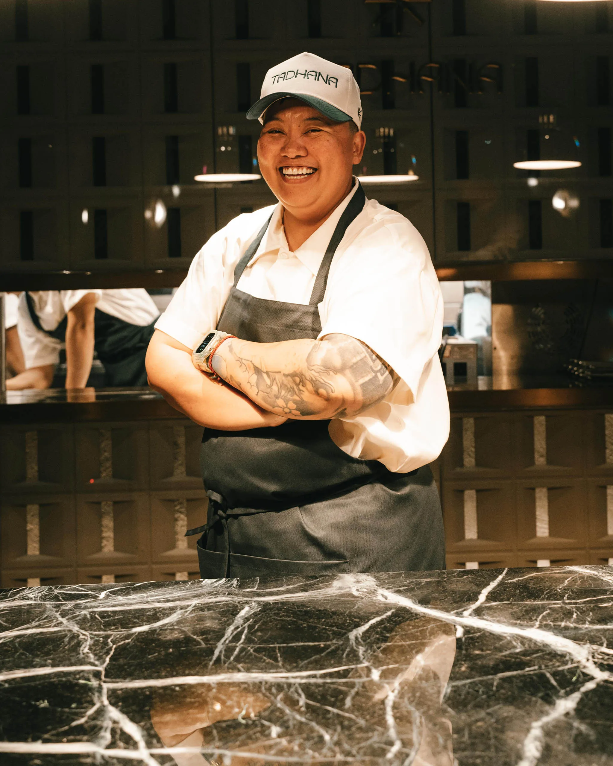 Smiling male chef in white shirt and gray apron stands behind a marble counter in a kitchen.