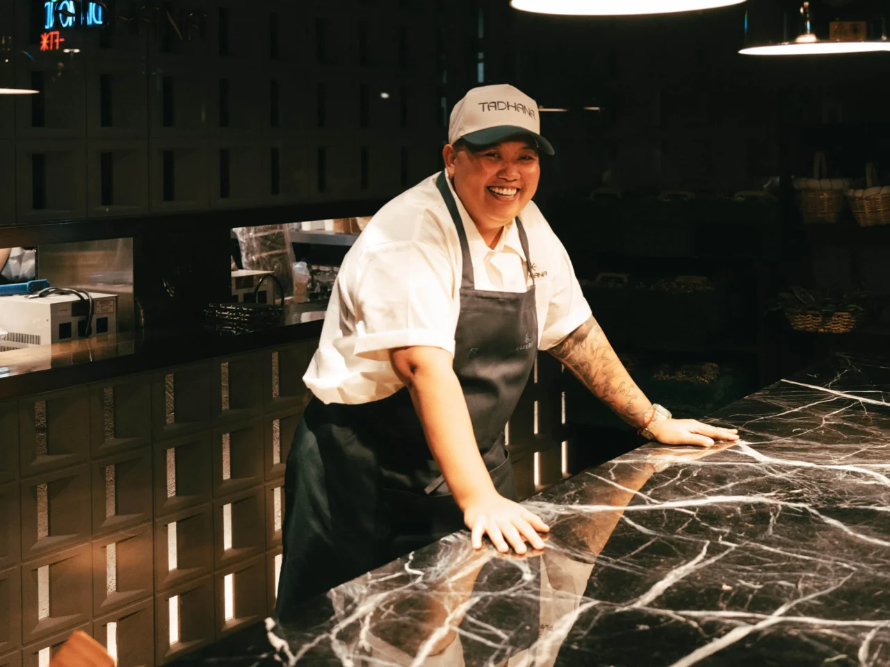 Group of restaurant staff in white shirts and green aprons posing behind a marble counter in a modern kitchen.