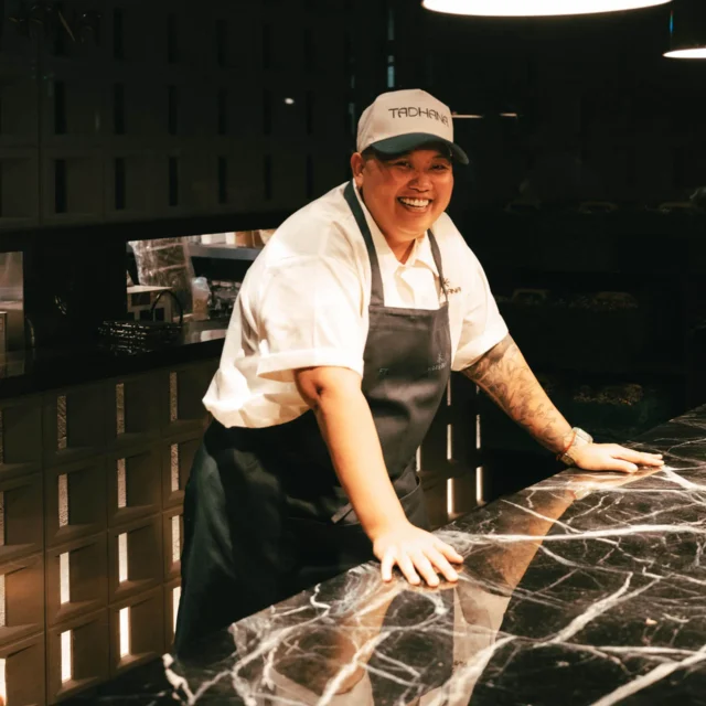 Chef smiling while leaning on a marble countertop in a dim kitchen, wearing a white shirt and dark apron with a white cap.