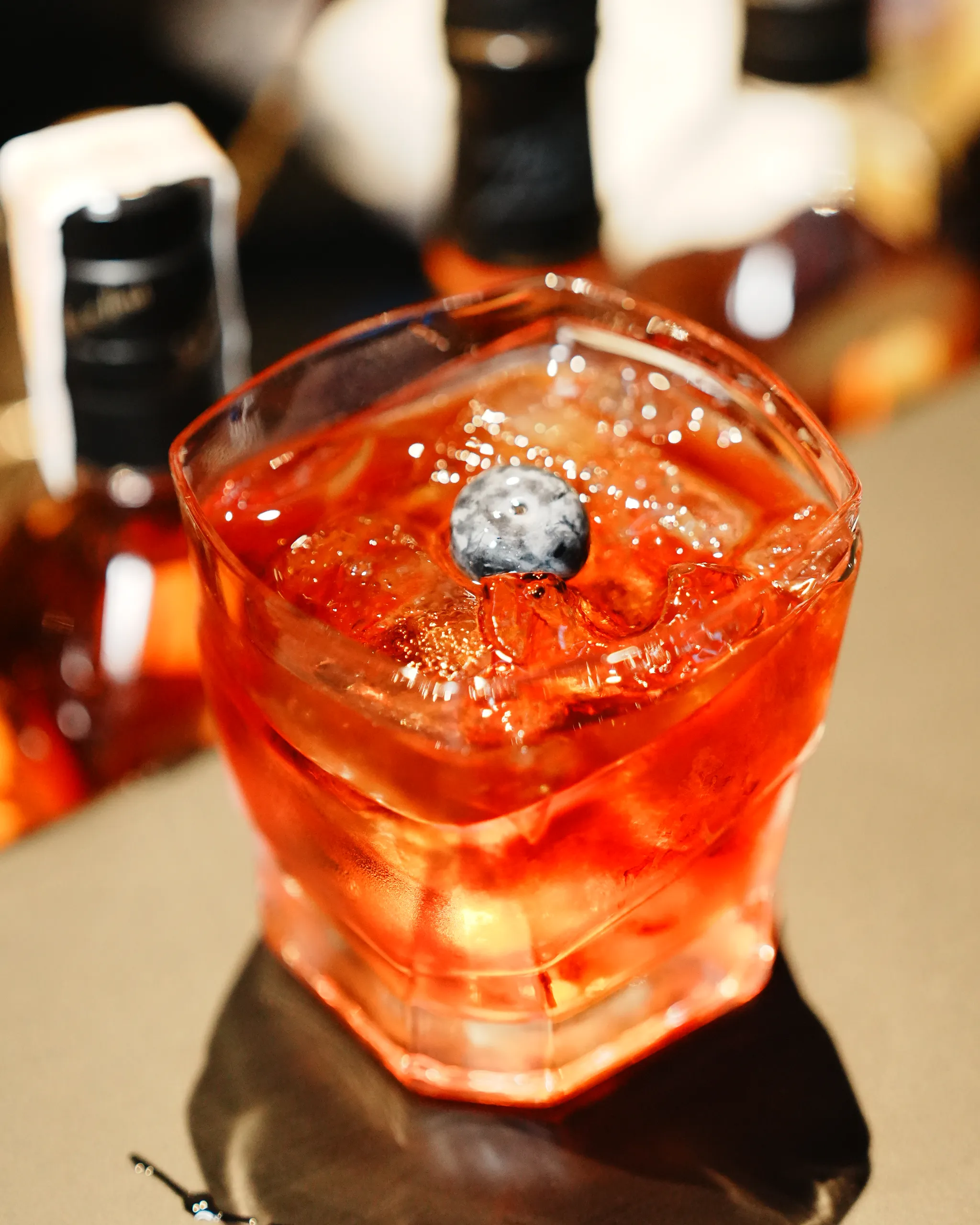 A close-up of a bright red cocktail with ice and a blueberry, in a crystal glass, blurred bar background behind it.