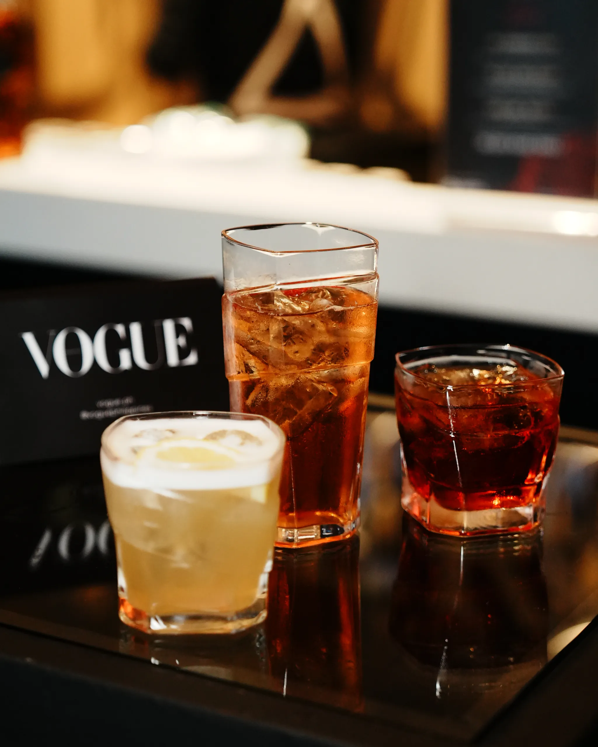 Tall, ice-filled amber cocktails on a glossy bar surface with a Vogue magazine backdrop in soft focus.