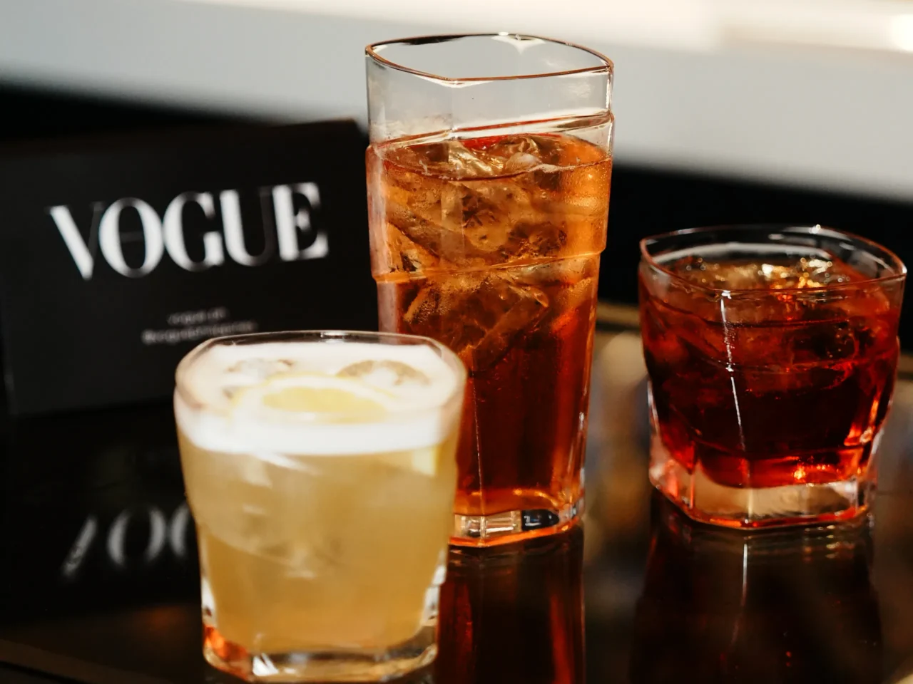 Three cocktails with ice on a reflective bar surface, a tall amber drink centered, and a blurred Vogue sign in the background.