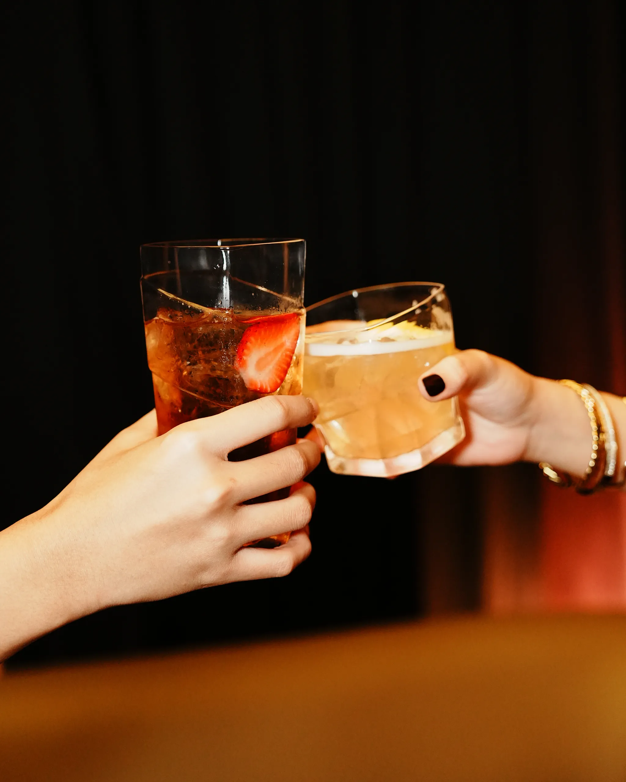 Two hands clinking glasses in a toast against a dark background, with strawberry and ice visible in the drinks.