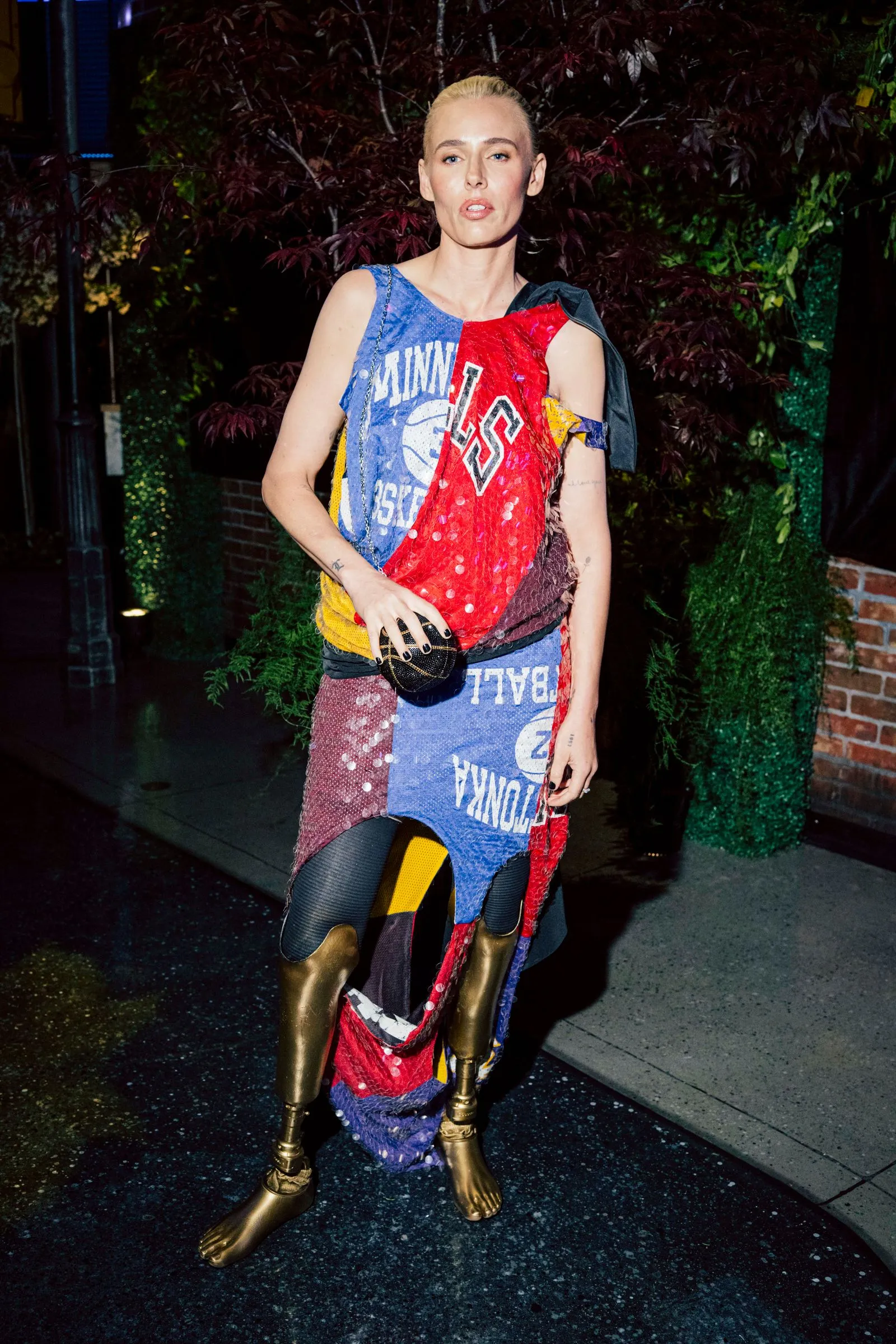 Person wearing a colorful patchwork dress made from sports jerseys, standing outdoors at night with leafy background.