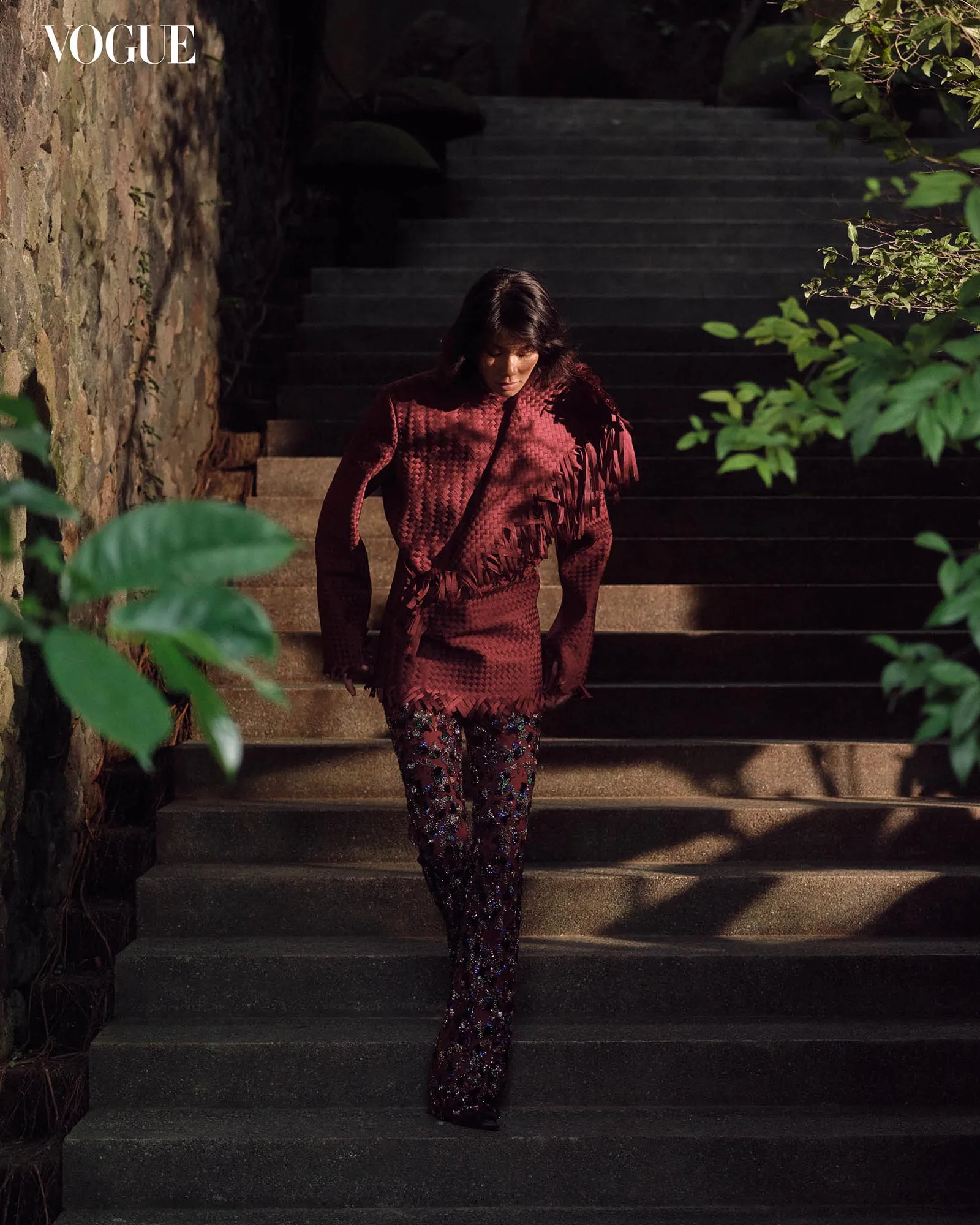 Editorial Vogue image: woman in burgundy textured knit with fringe and sequined pants walking down stone steps, with ivy and greenery nearby.