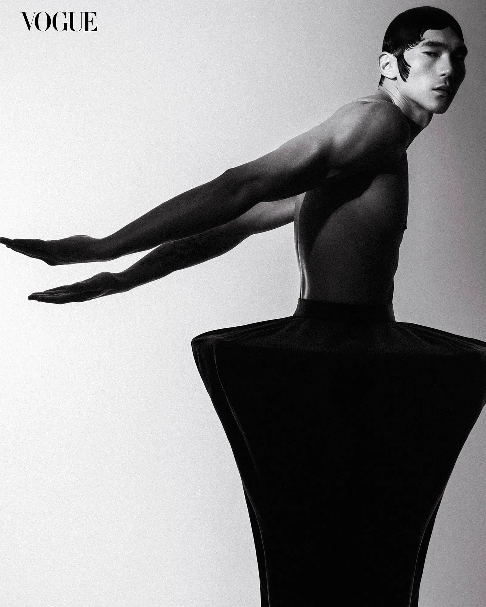 Black-and-white Vogue cover-style photo of a male ballet dancer in a tutu, arms extended outward across the frame.