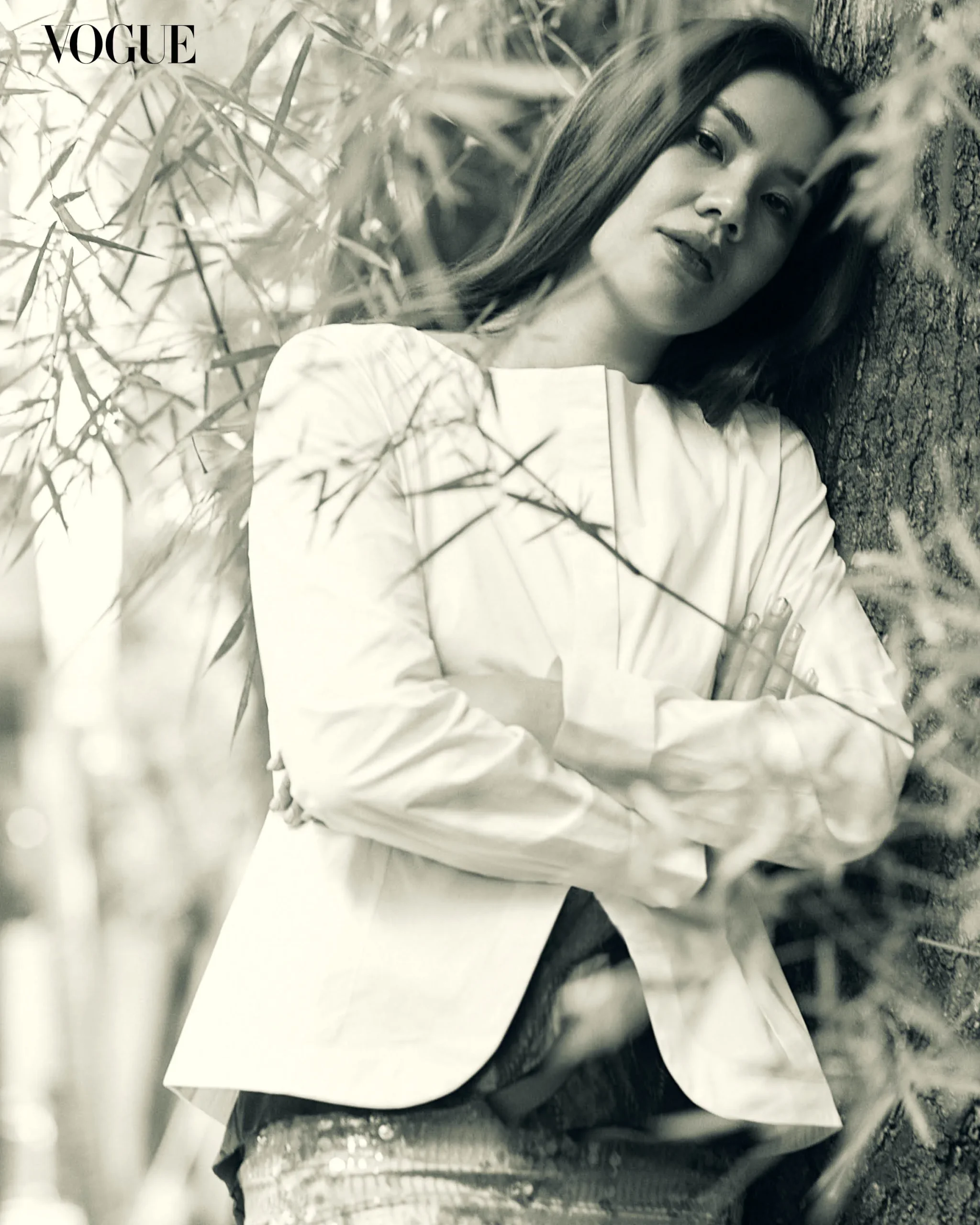 Black-and-white Vogue fashion photo of a woman with long hair leaning against a tree in a white blouse, arms crossed.