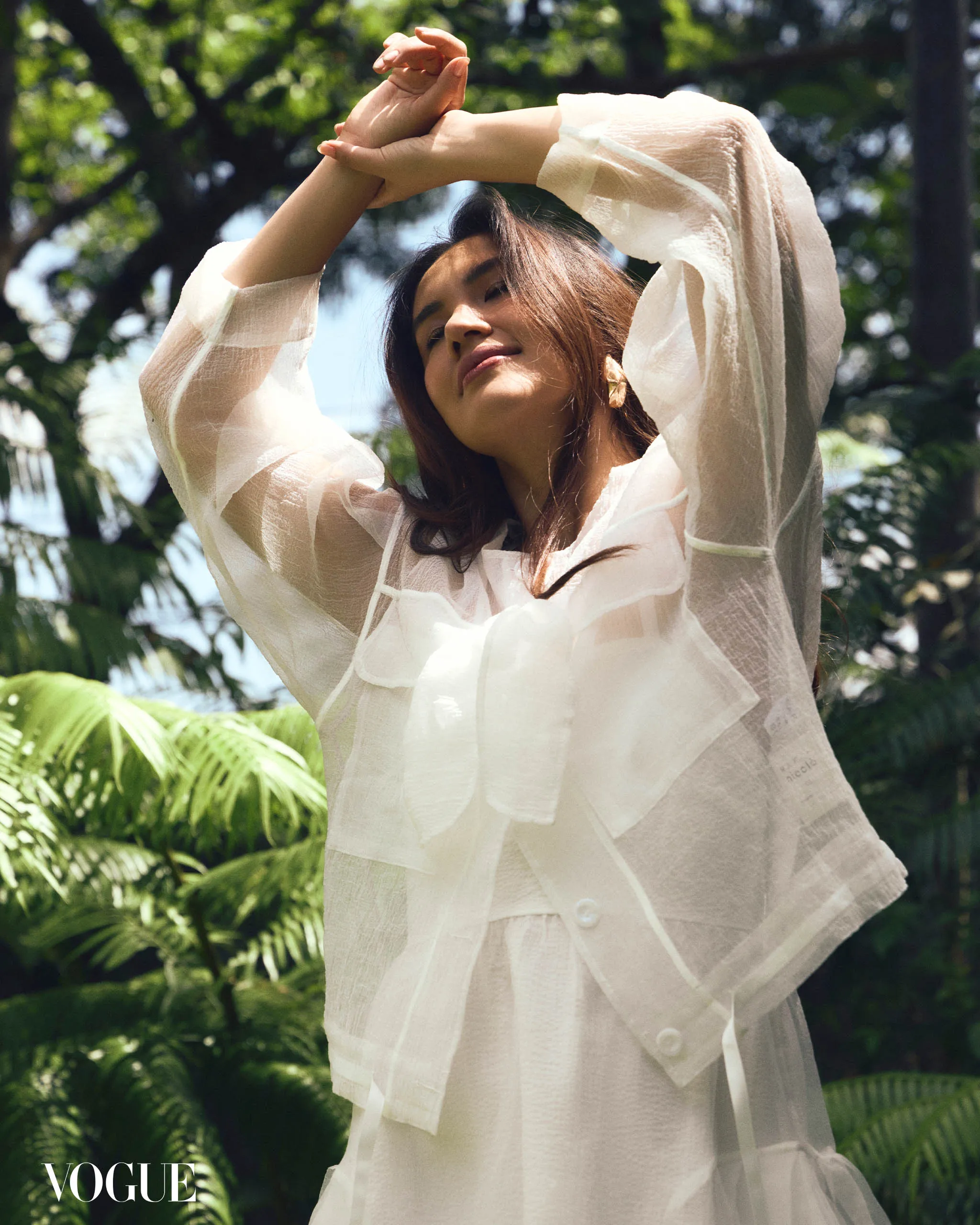 Woman in a sheer white blouse raises her arms outdoors, with greenery in the background (Vogue logo visible).
