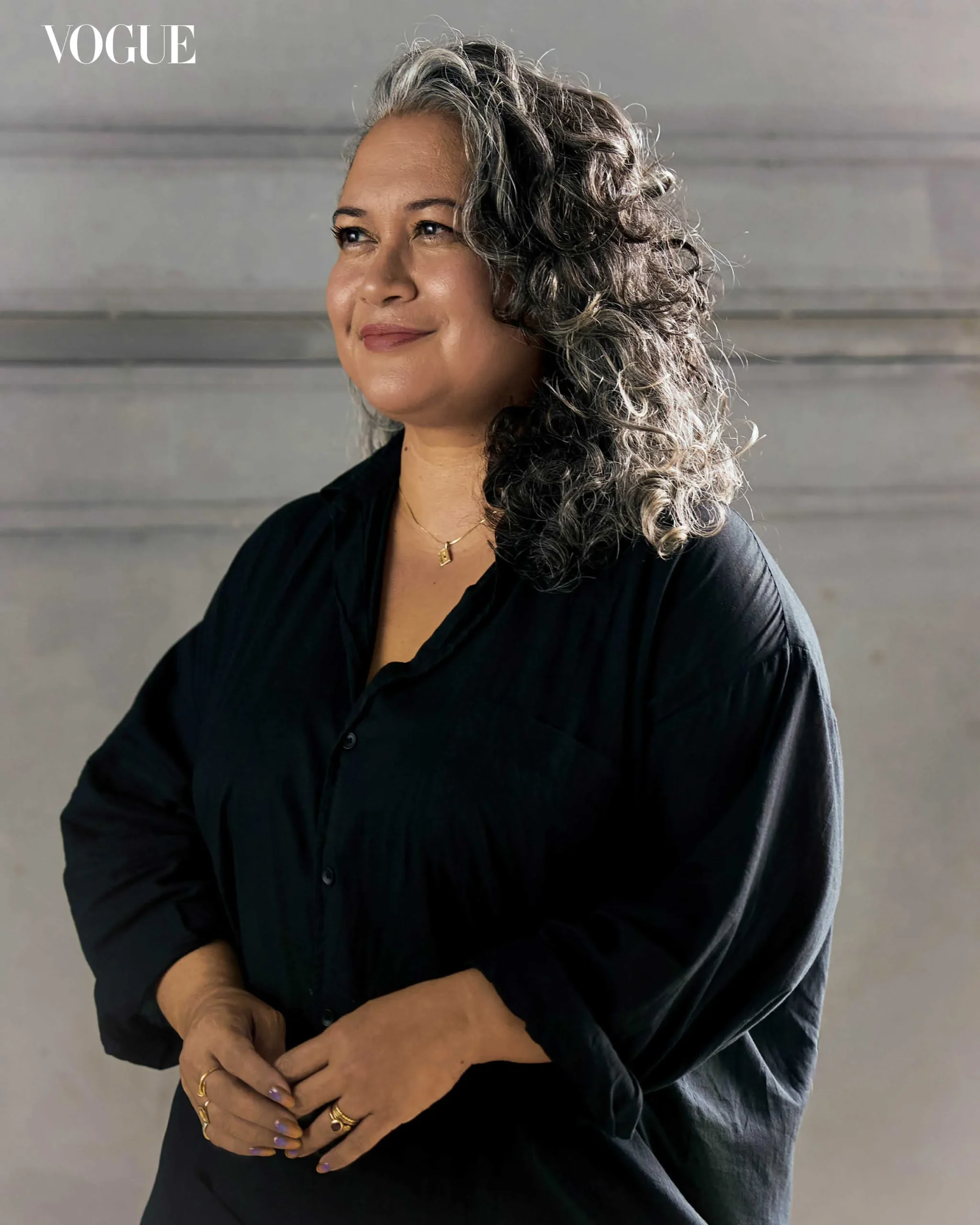 Portrait of a smiling woman with salt-and-pepper curly hair in a black shirt, standing against a gray wall with a Vogue logo in the top-left corner.
