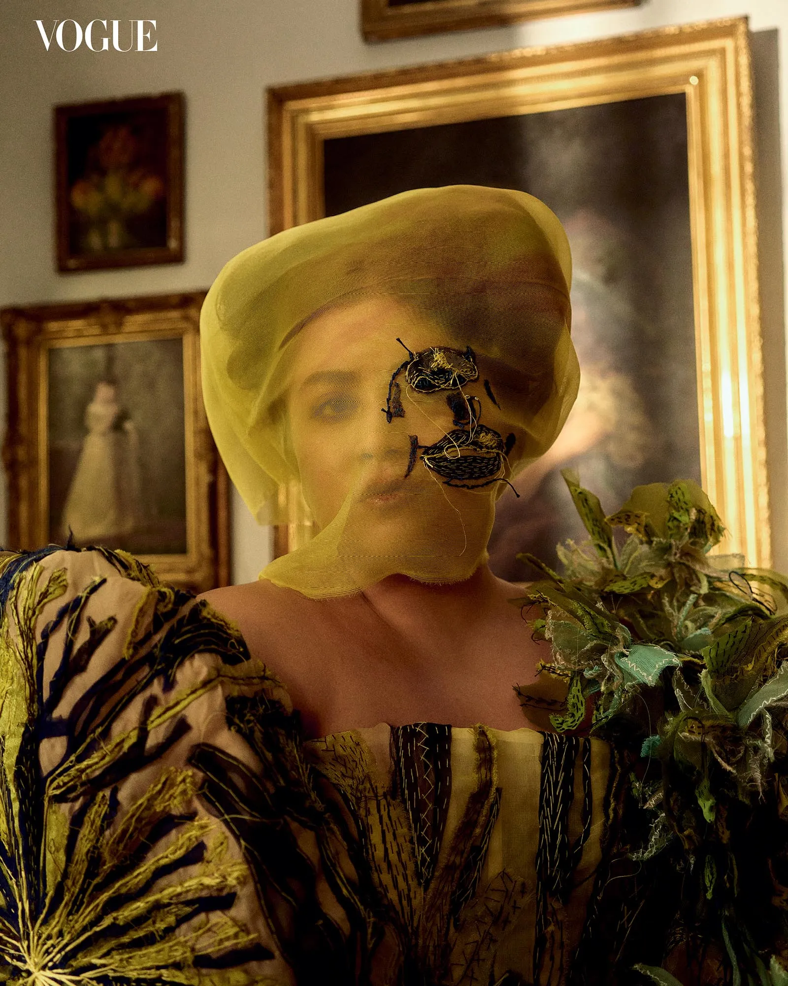 Model wearing a pale yellow veil over her face with live-looking insects attached, in a gallery with framed paintings; Vogue logo visible.
