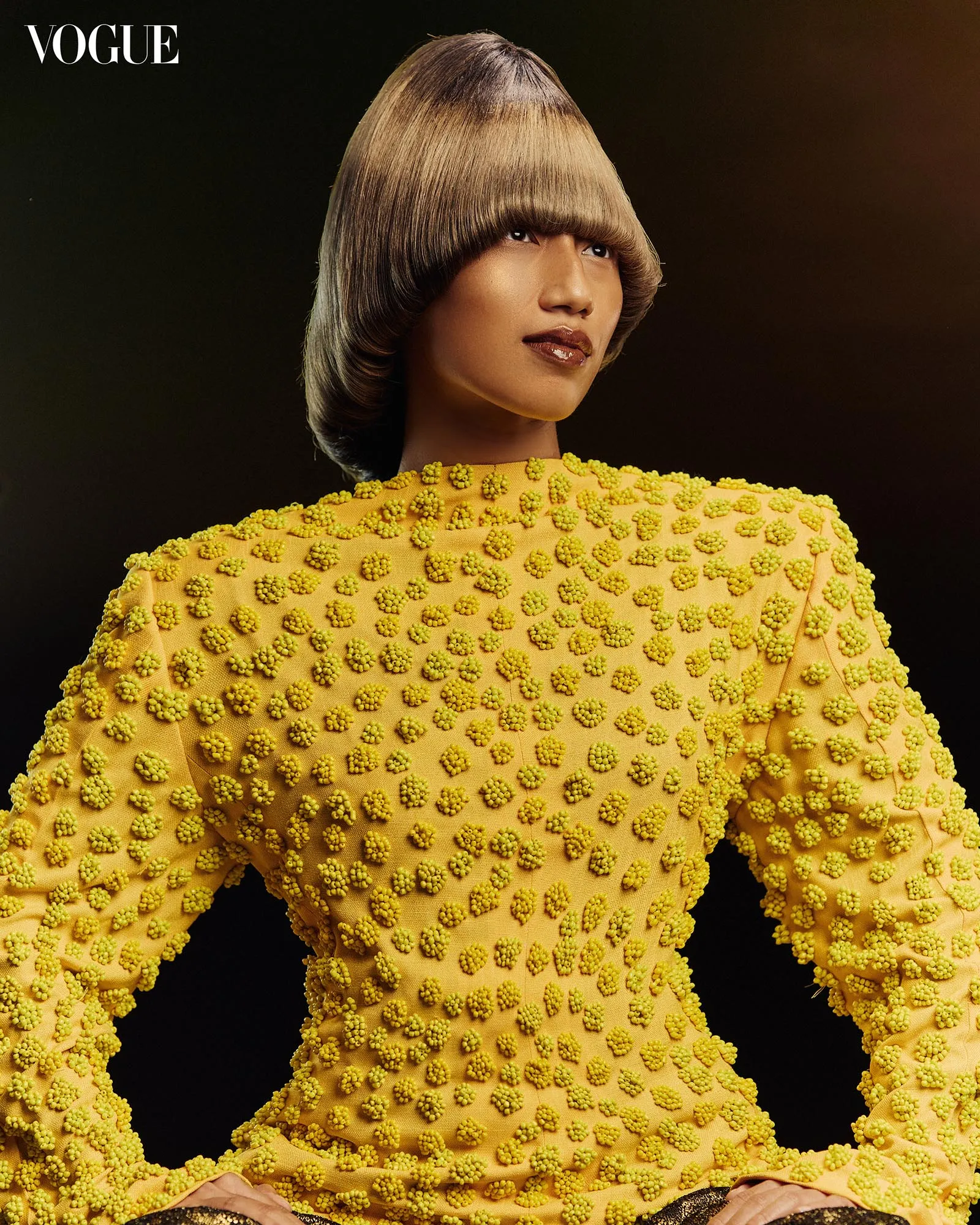 Model wearing a bright yellow dress densely covered with small pom-poms, against a dark backdrop (Vogue). They pose with hands visible at waist level.