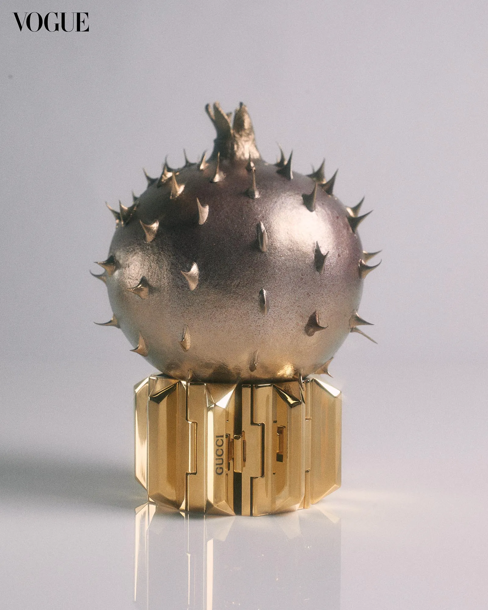 Close-up of a gold Gucci perfume bottle with a spiked, spherical cap against a pale background, Vogue logo visible in the corner.