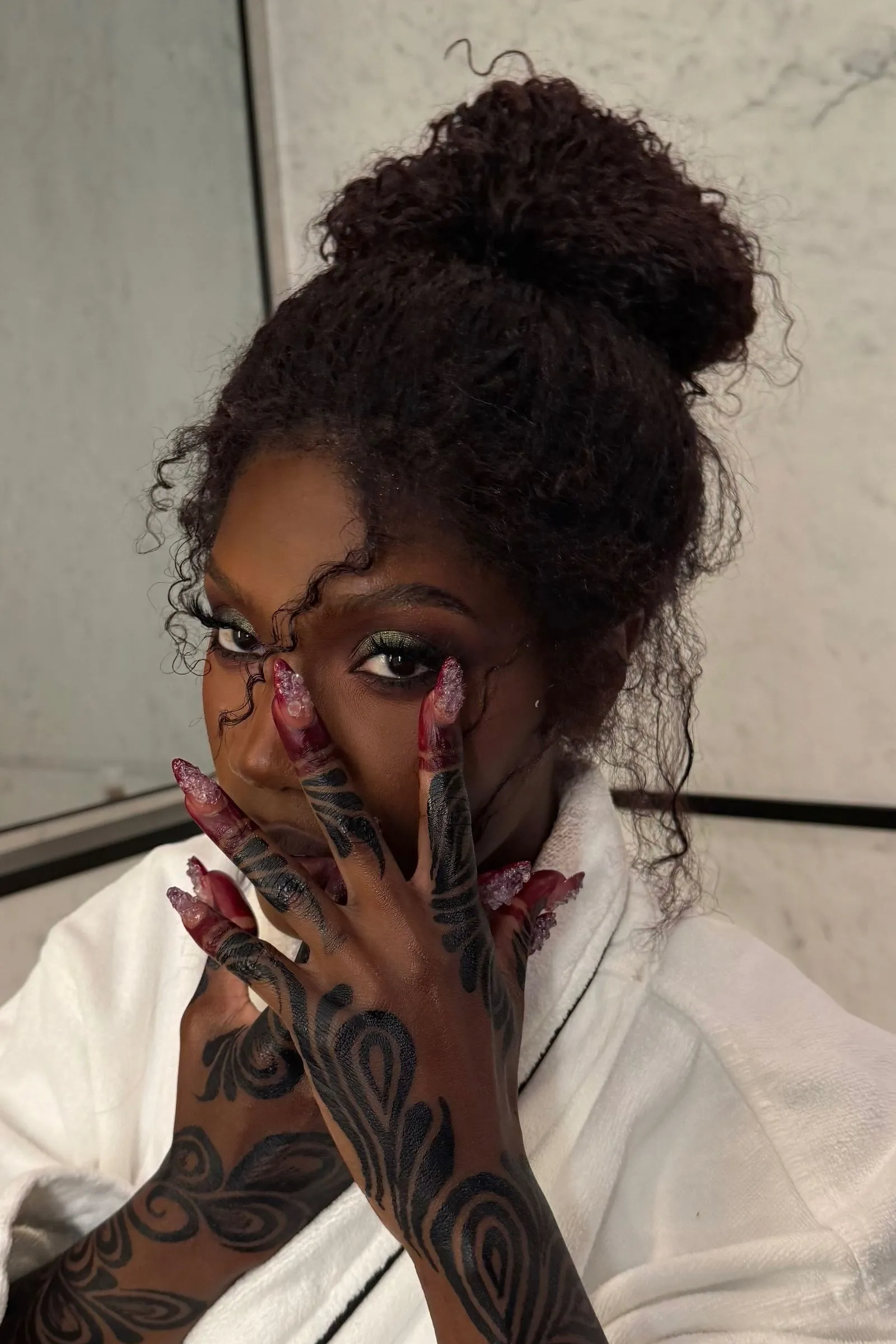 Close-up of a person with a dark topknot bun, wearing a white robe, showing long red glitter nails and black tattooed sleeves across the face/hand area.