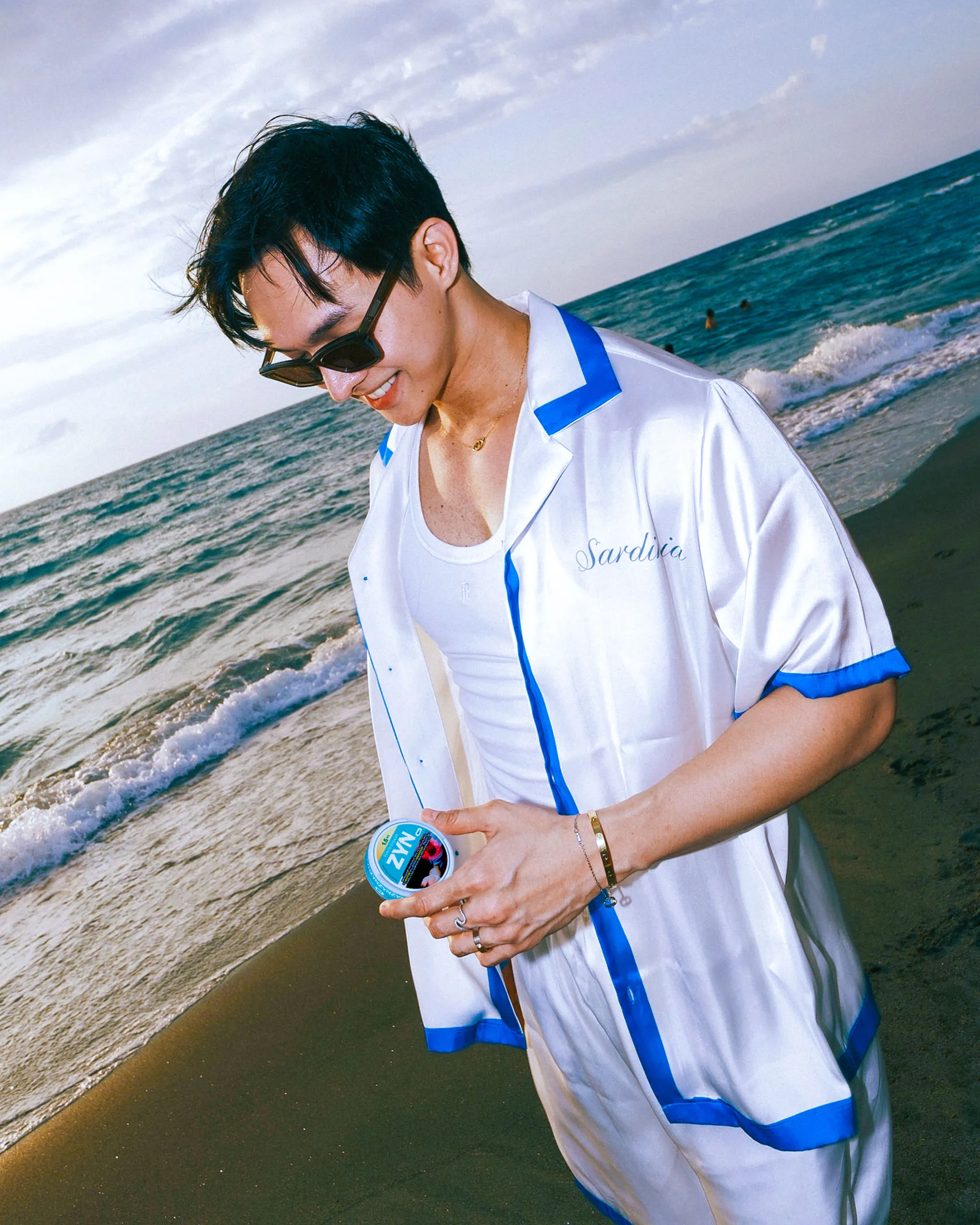 Person in a white and blue beach outfit wearing sunglasses, standing on the shore with waves behind. (informative)