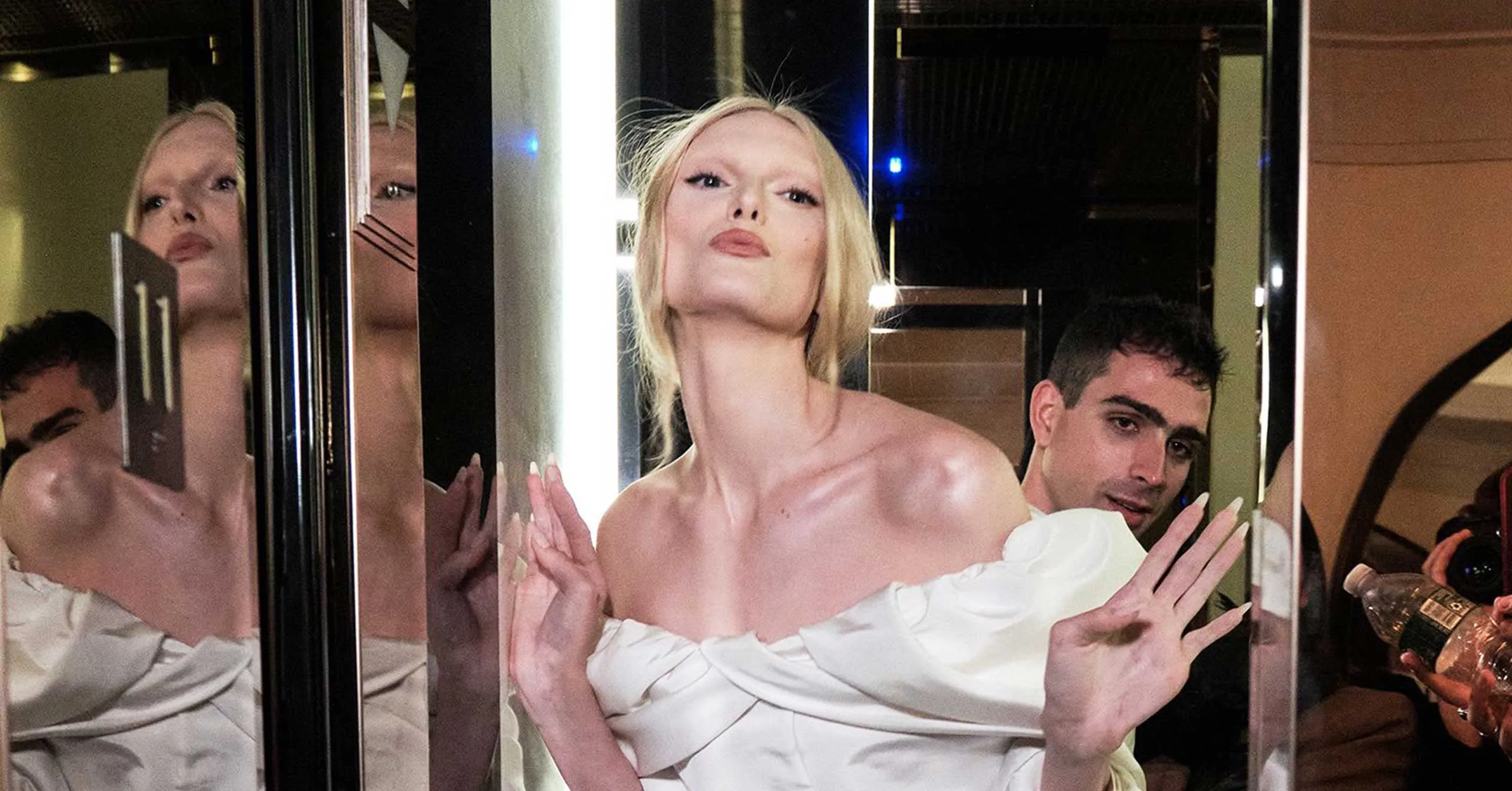 Blonde woman in an off-shoulder white dress posing against mirrored panels at a nightclub, with two men nearby and party lighting in the background.