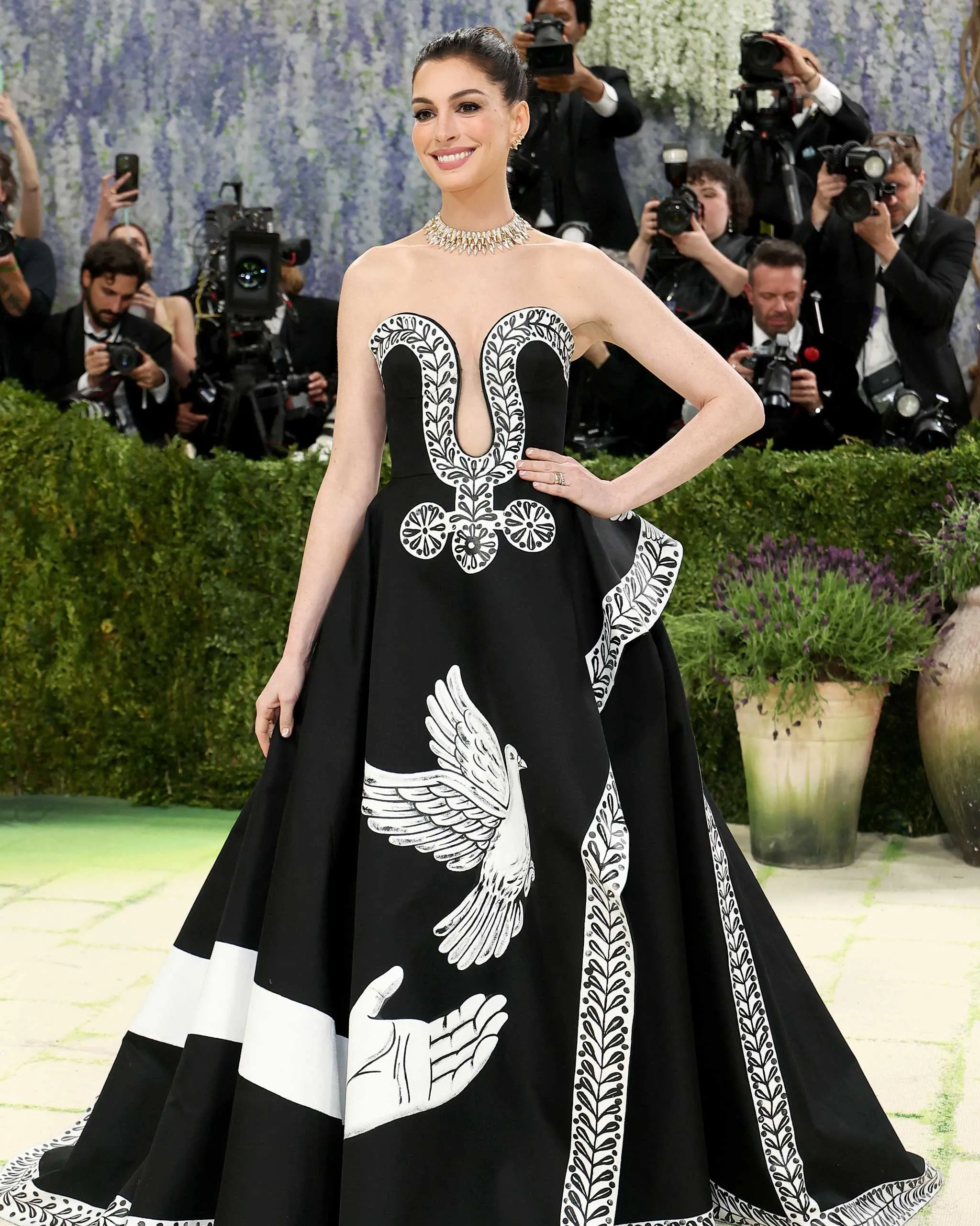 Woman on a red carpet wearing a black gown with white embroidered pattern, smiling at the cameras with photographers in the background.
