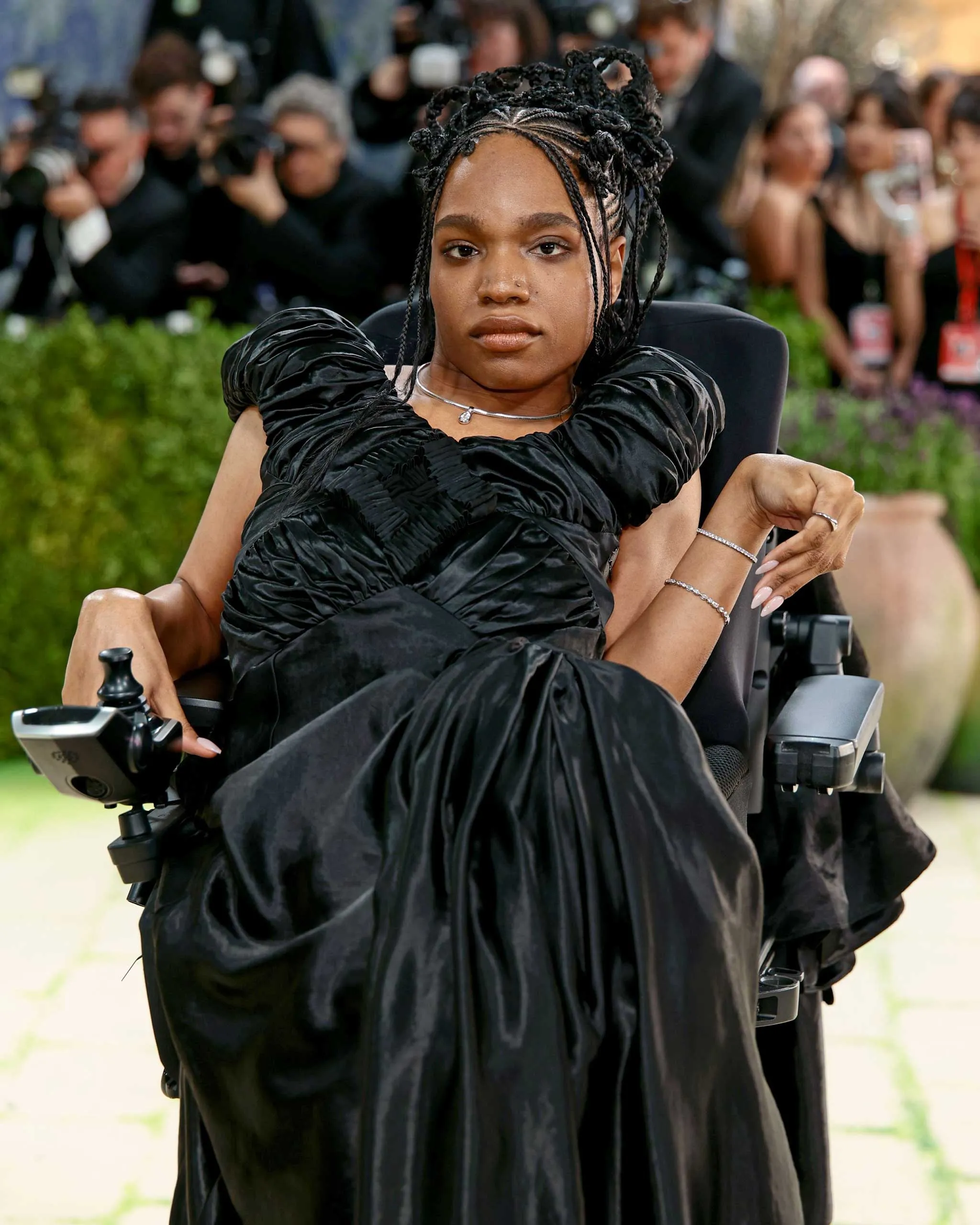 Woman in a black formal gown seated in a wheelchair, with photographers blurred in the background at a crowd event.