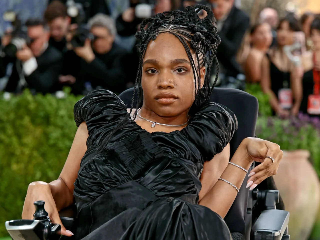Woman with braided updo wearing a black ruffled dress poses on a red carpet, photographers in the background.