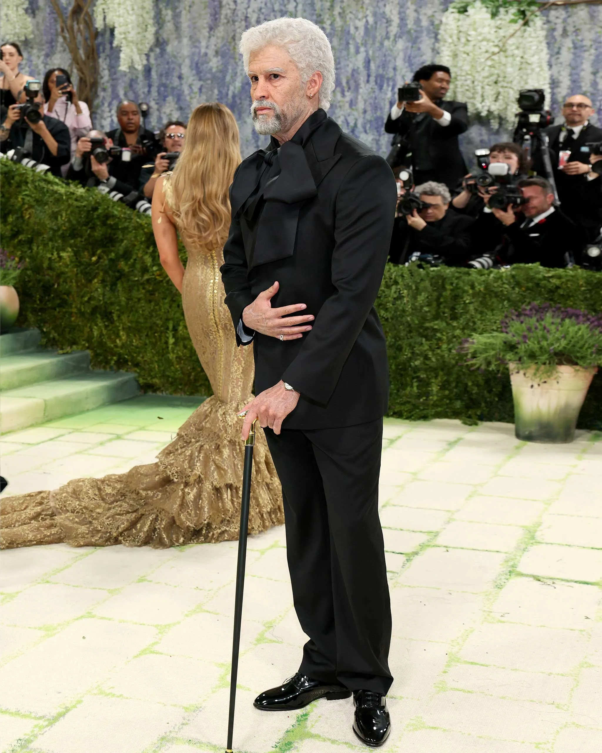Man with white hair and gray beard in a black tuxedo posing on a photo gallery backdrop, surrounded by photographers and cameras.