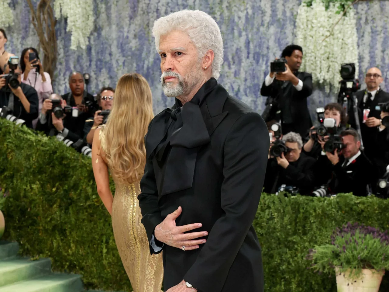 Man with white hair and beard in a black suit on a red carpet, surrounded by photographers and flashing cameras.