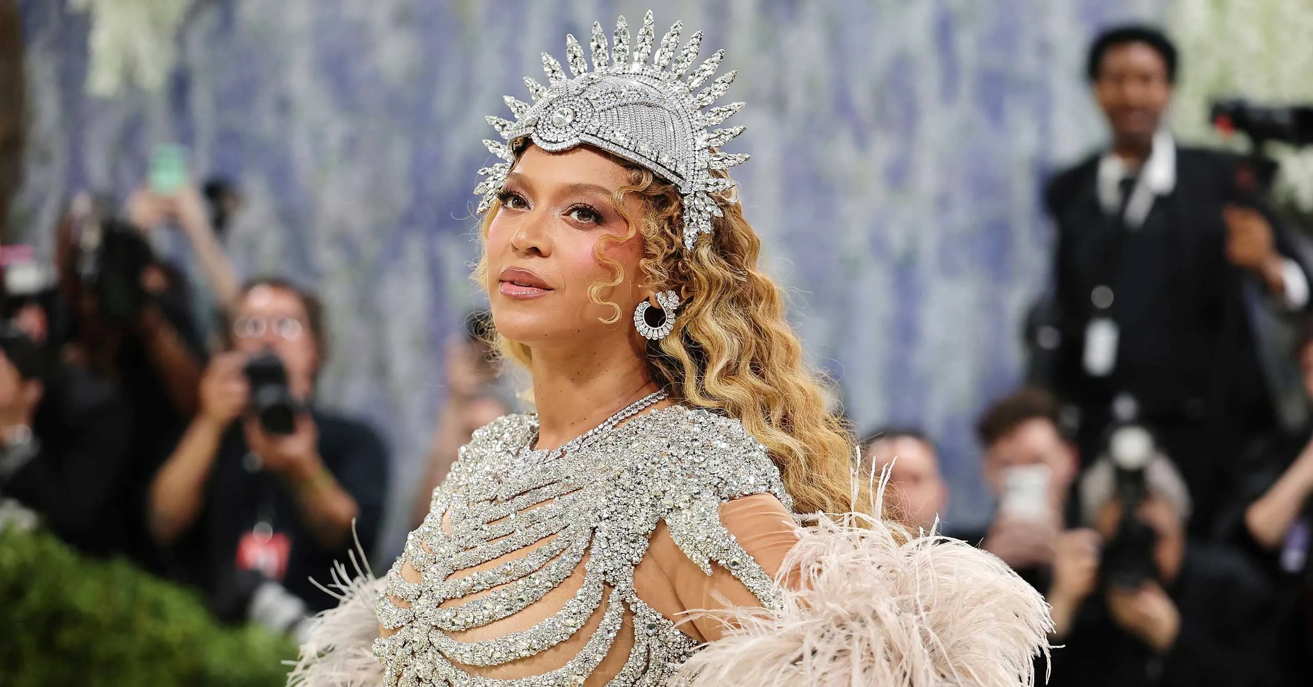 Woman on a red carpet wearing a silver, diamond-studded headdress and matching sparkling gown, surrounded by photographers.