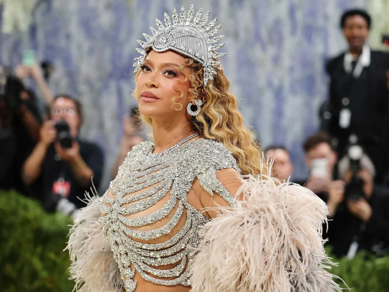 Woman on a red carpet wearing a silver, diamond-studded headdress and matching sparkling gown, surrounded by photographers.