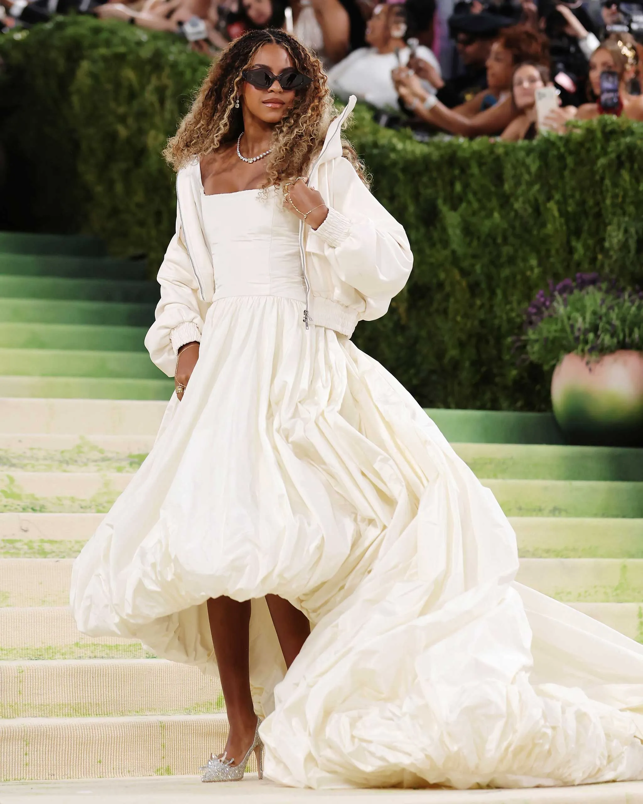 Woman in a white strapless gown with a voluminous, crinkled skirt, wearing sunglasses and a pearl necklace on steps as photographers snap photos.