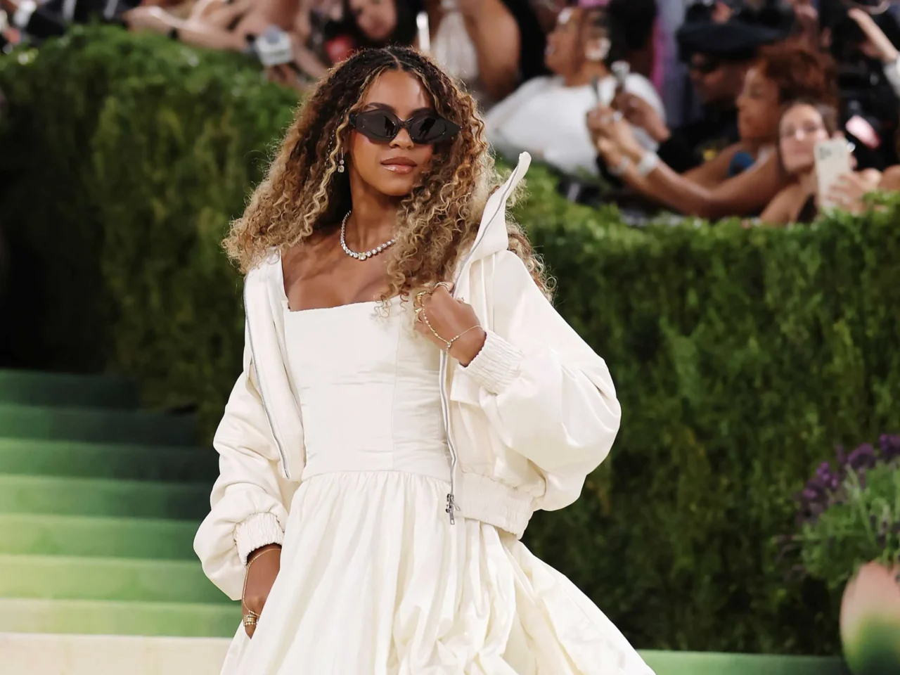 Stylish woman in a white outfit and dark sunglasses posing for photographers as fans crowd the background at a public event. (Celebrity on a stepped green carpet)