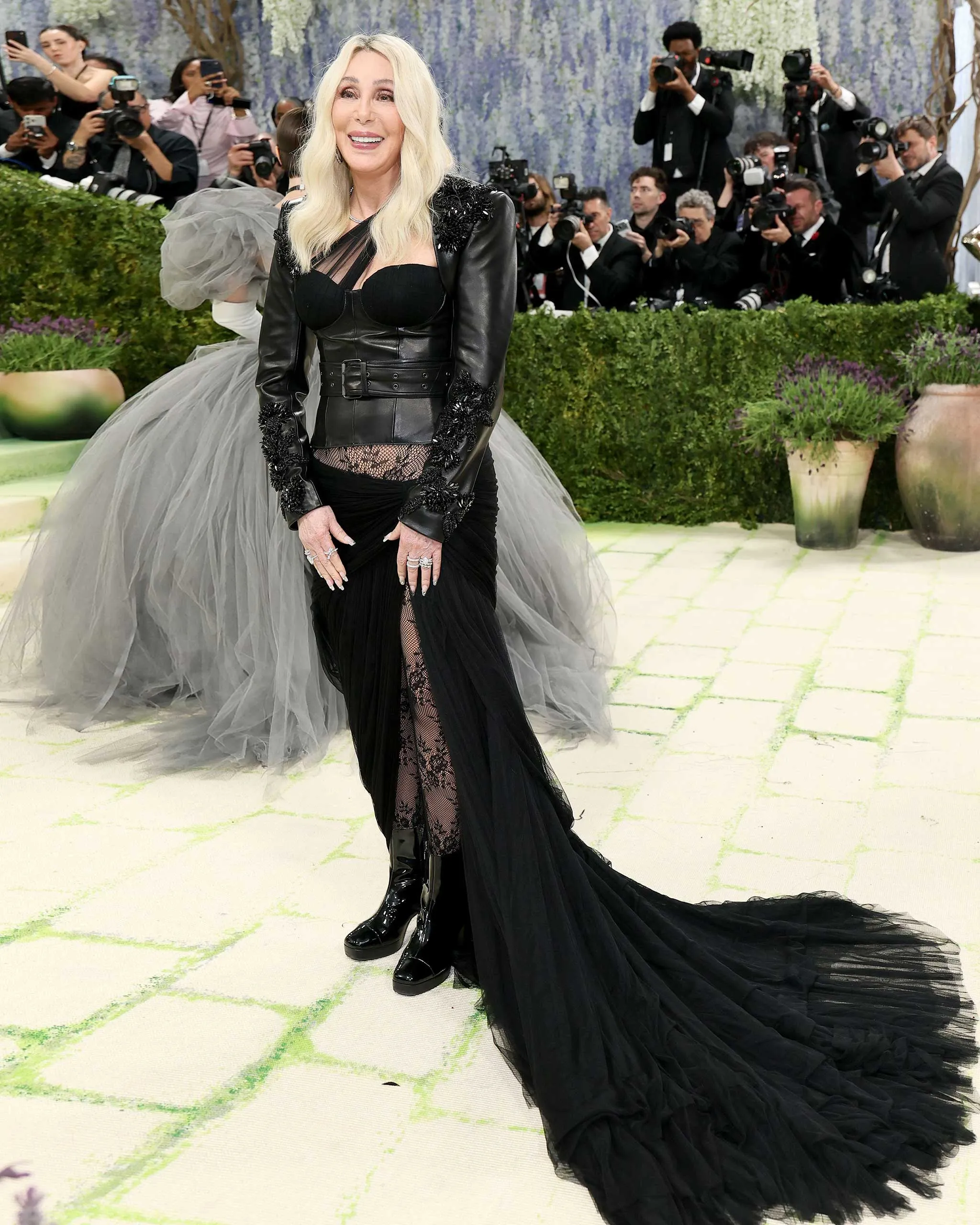 Woman in a black leather-paneled gown with lace details and a long sheer train posing as photographers crowd behind her.