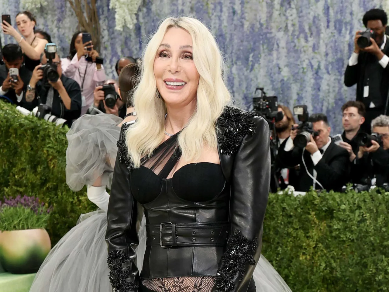 Blonde woman in a black outfit smiles on a red carpet, surrounded by paparazzi and photographers.