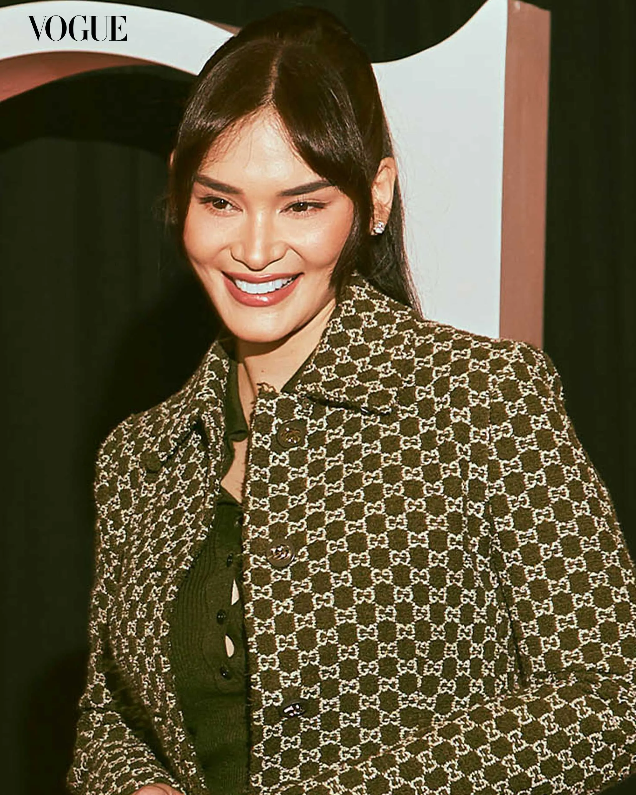 Smiling woman wearing a brown Gucci-patterned coat at a Vogue event; Vogue watermark visible in the corner.