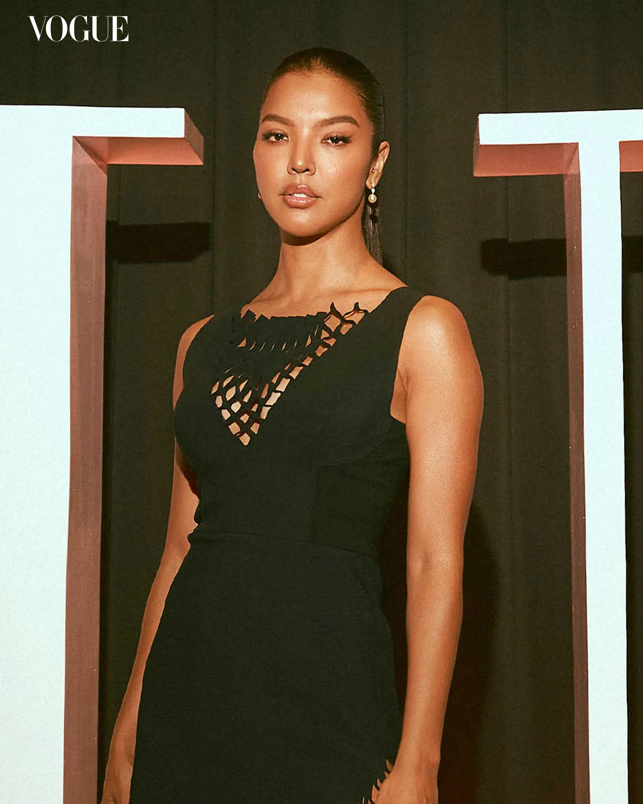 Model wearing a black sleeveless dress with a lattice-cut neckline, posing against a dark backdrop with pale teal framed panels; Vogue logo in the corner.