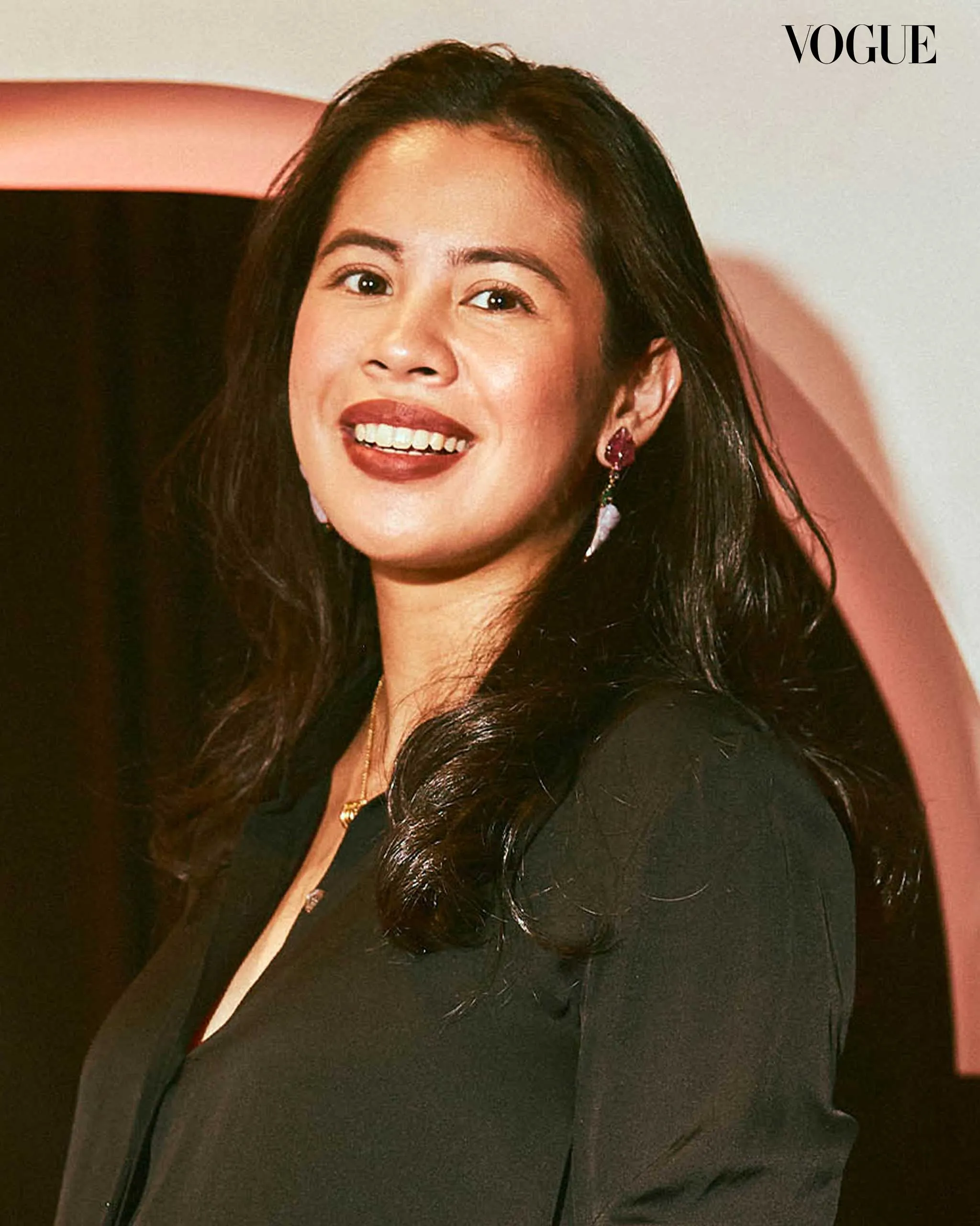 Close-up portrait of a smiling woman with long dark hair wearing a black blazer and pink earrings; Vogue logo in the corner.