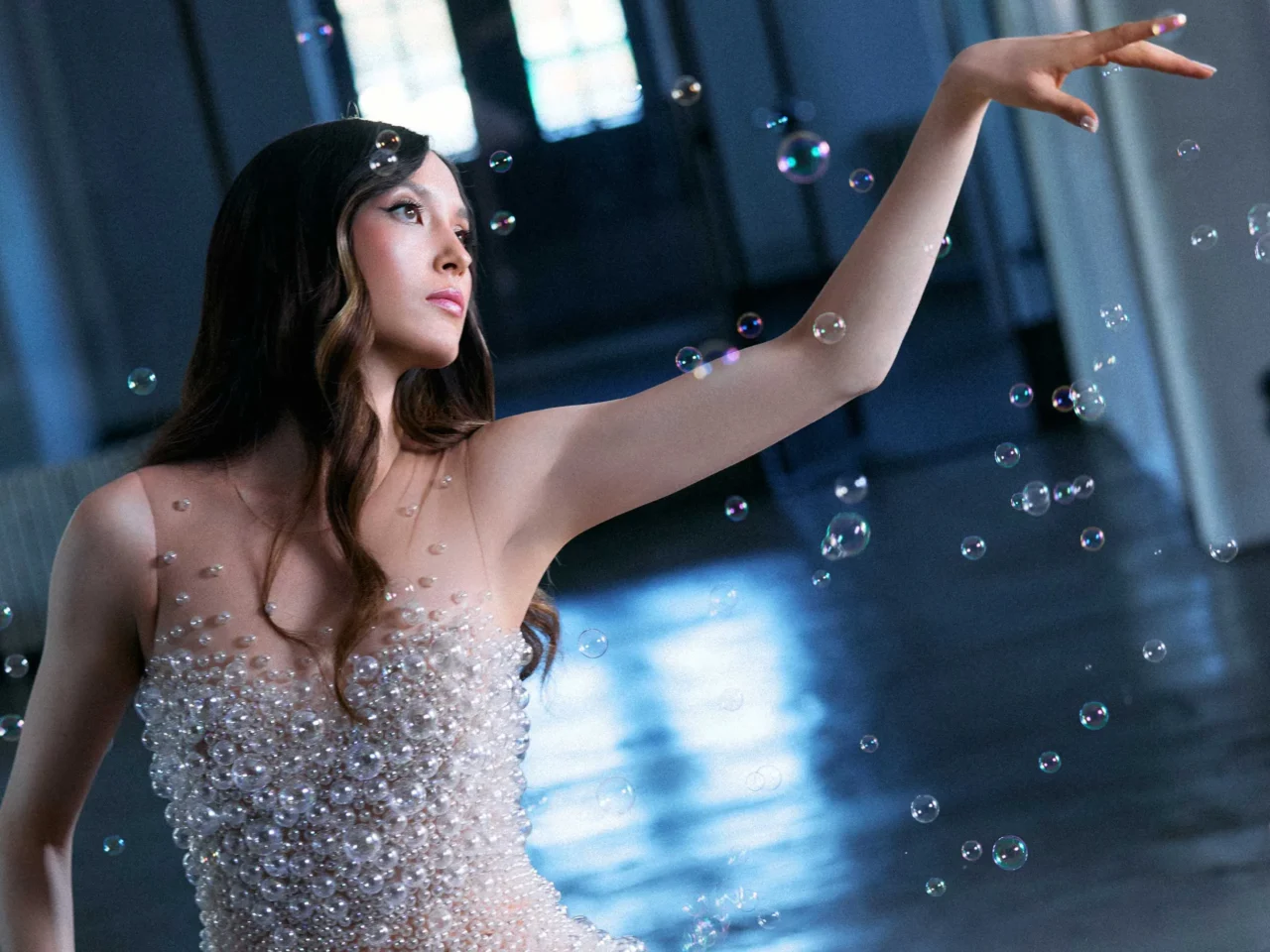 Woman gracefully reaches upward among floating bubbles, wearing a beaded, strapless dress in a blue-toned setting.