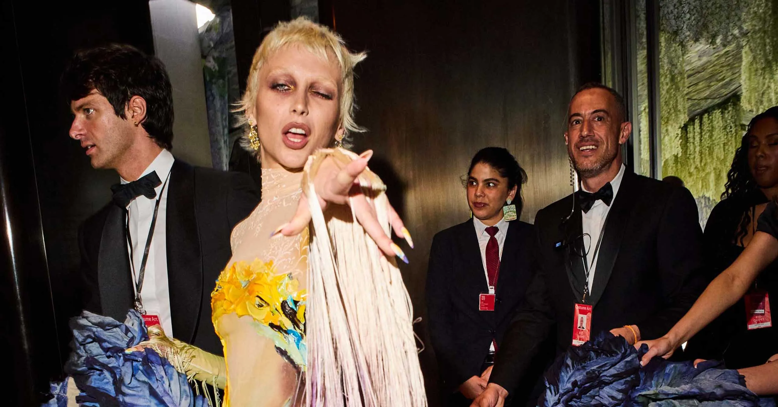 Center-stage blonde woman in a beige fringed dress points at the camera while surrounded by suited staff with lanyards on backstage at a fashion show.