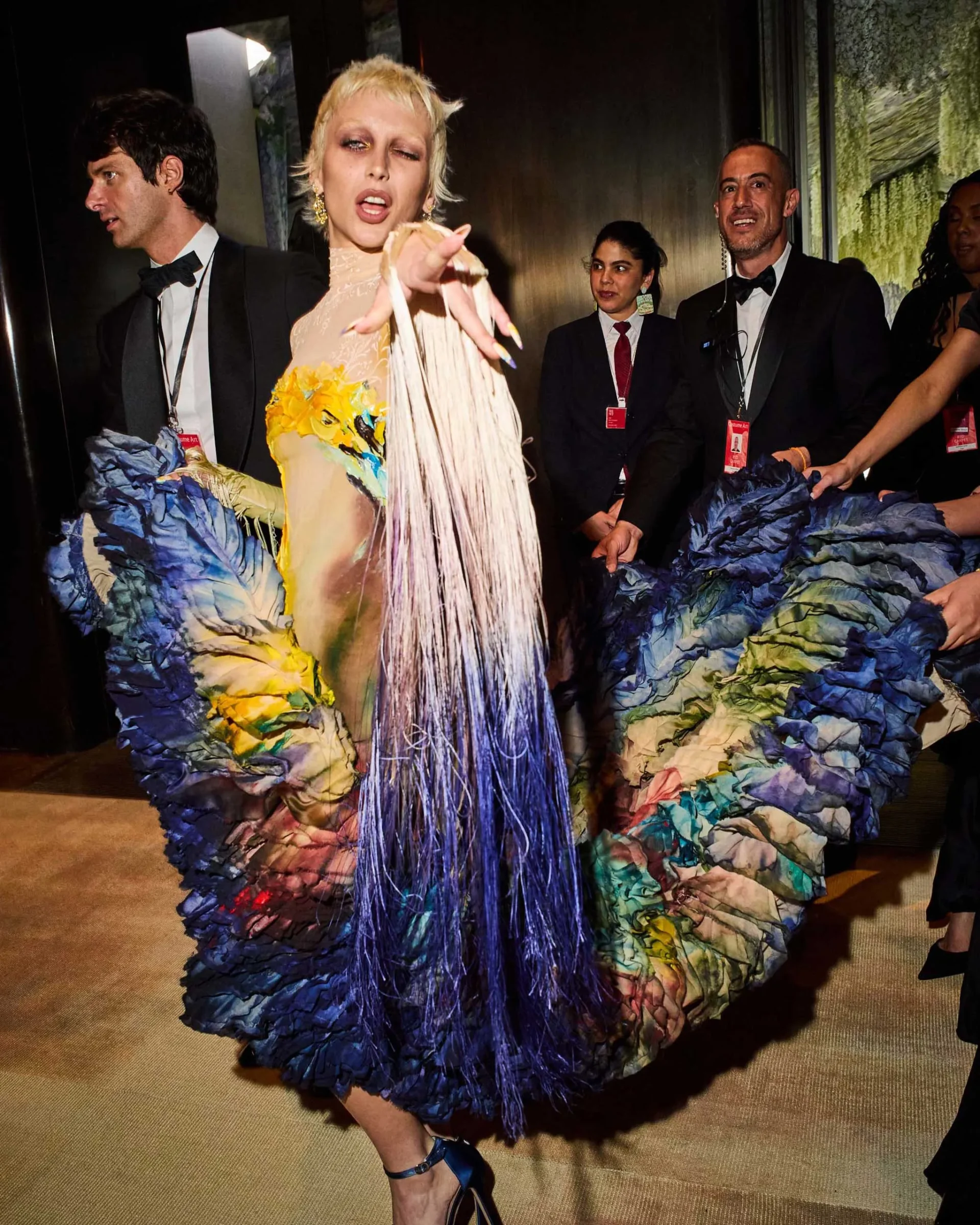 Center-stage blonde woman in a beige fringed dress points at the camera while surrounded by suited staff with lanyards on backstage at a fashion show.